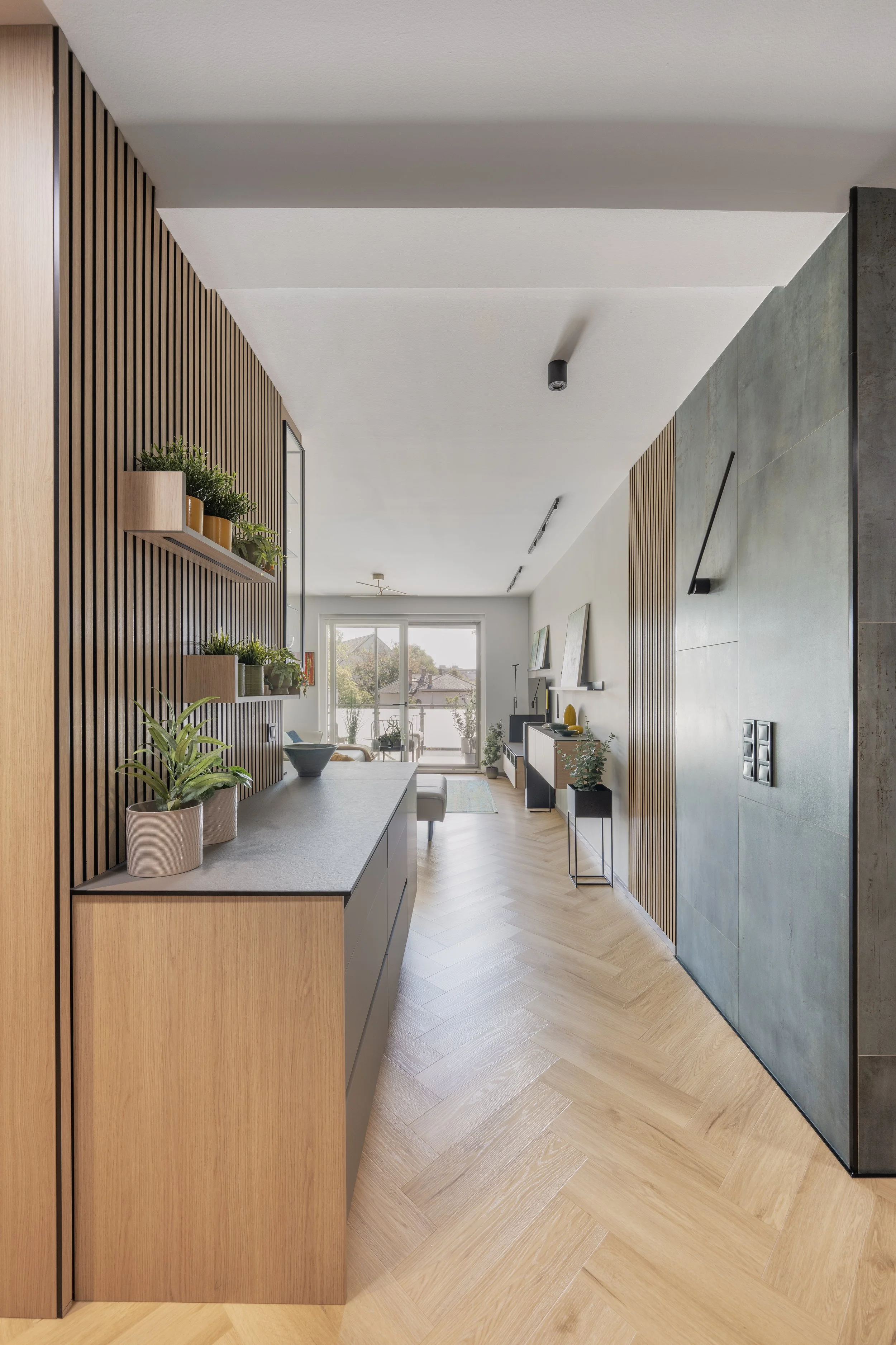 Modern living space with wooden vertical slats on the wall, shelves with potted plants, a gray countertop, and a view of a balcony with outdoor furniture and trees through large windows.