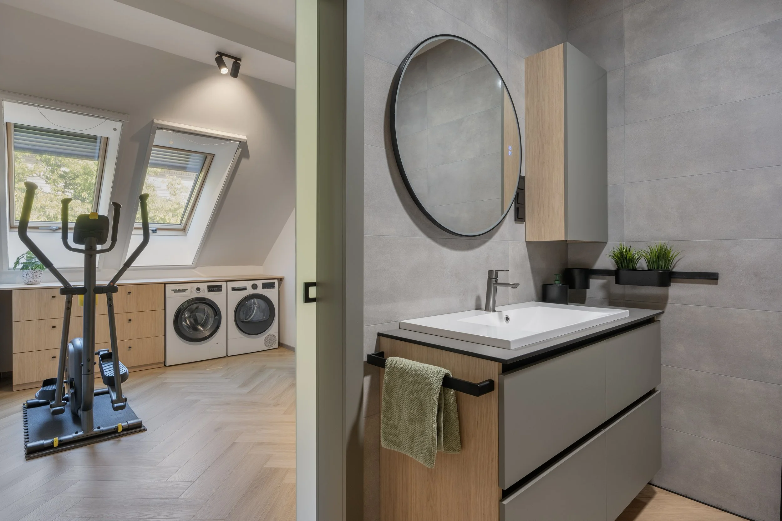 Split view of a laundry room and a bathroom. The laundry room has two slanted windows, light wood cabinets, a black exercise machine, and laundry appliances. The bathroom has a round mirror, wooden and gray cabinets, a sink, a small black shelf with green plants, and gray tiled walls.