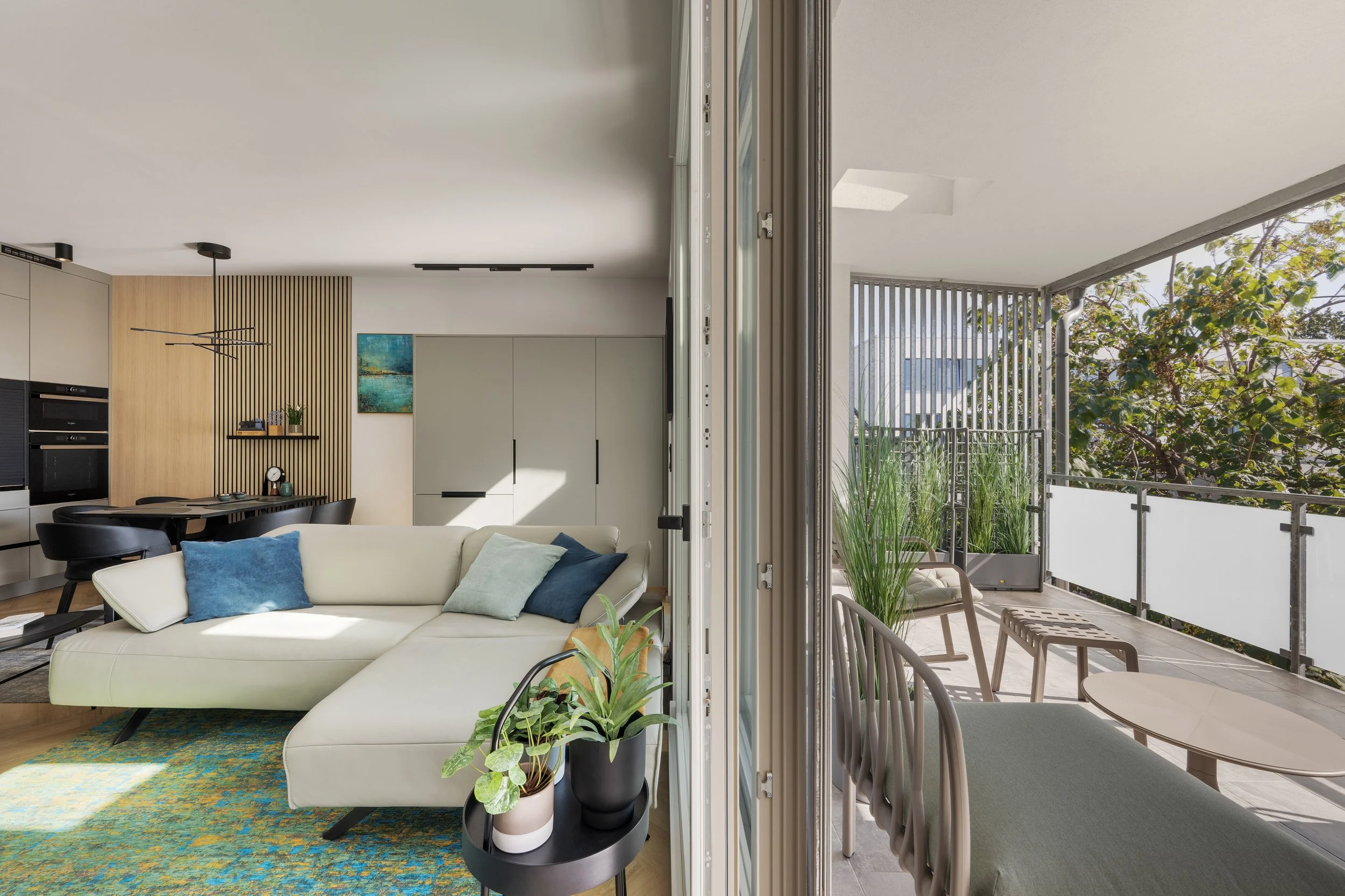 View of a modern living room connected to a balcony, featuring a white sofa with blue and green pillows, wooden flooring, a patterned rug, and a sliding glass door opening to a balcony with outdoor seating, plants, and a view of trees.
