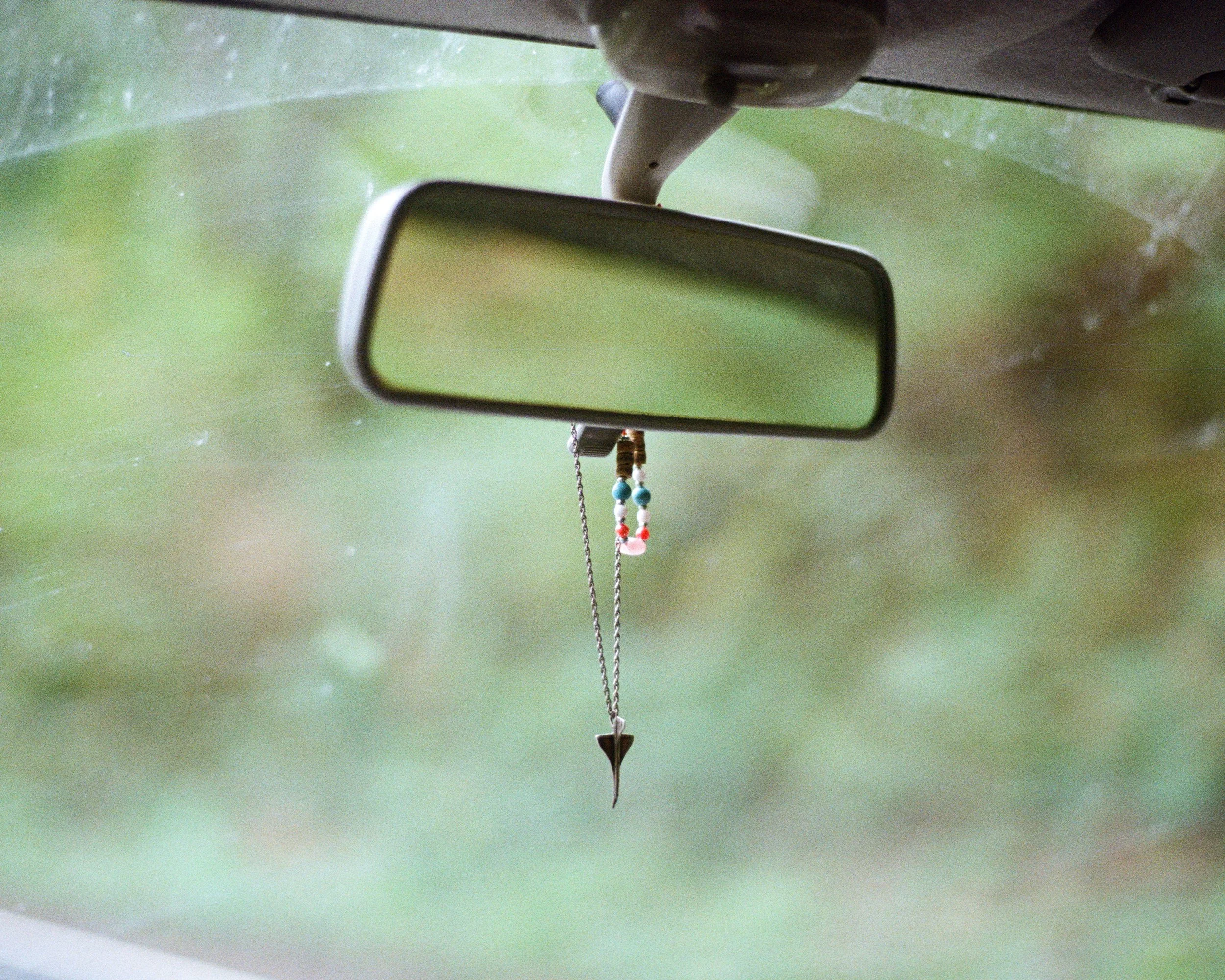 Car's interior rearview mirror with a beaded necklace hanging from it, and a green blurry background seen through the windshield.