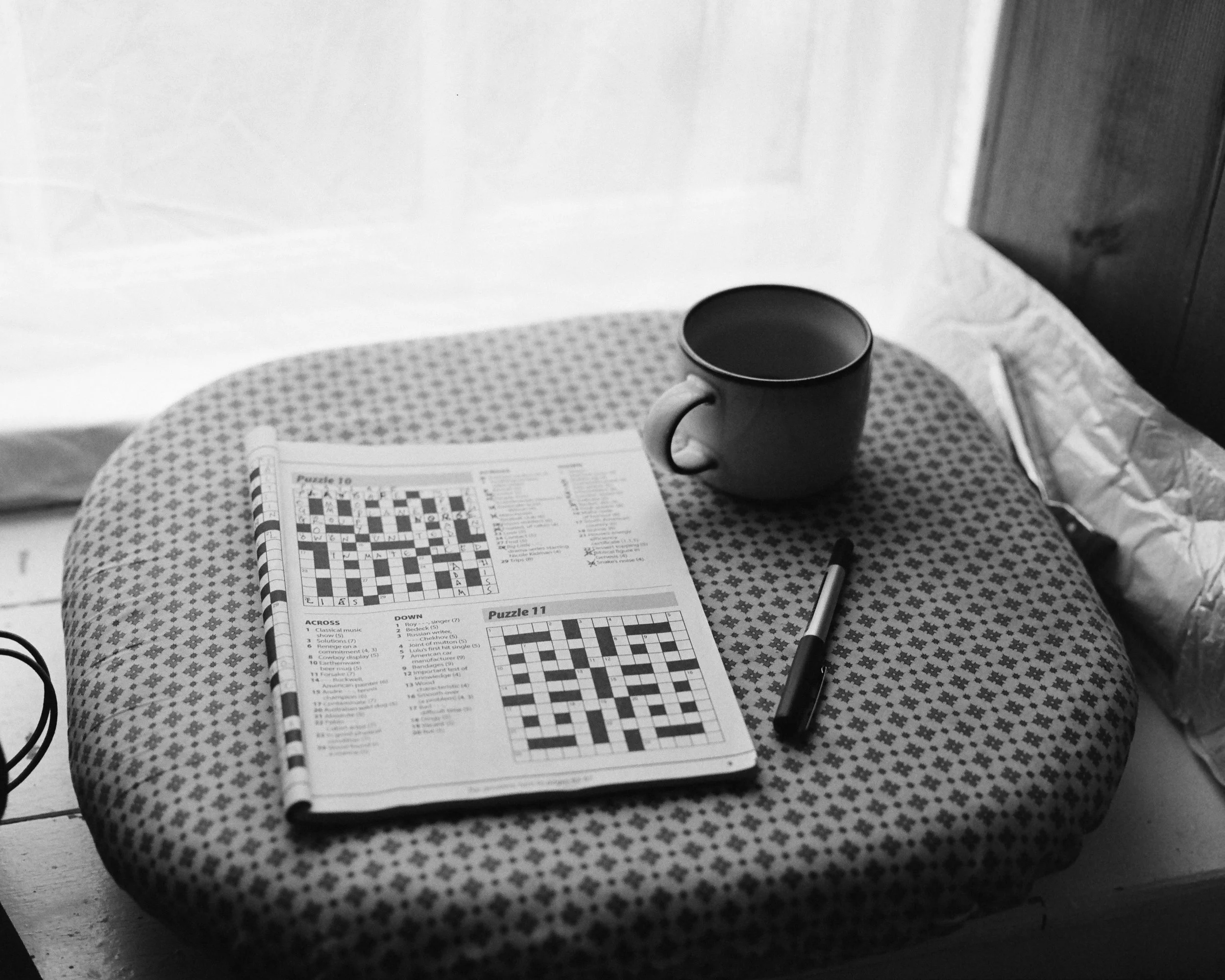 Black and white photo of a crossword puzzle book, pen, and mug on a patterned cushioned surface near a window.