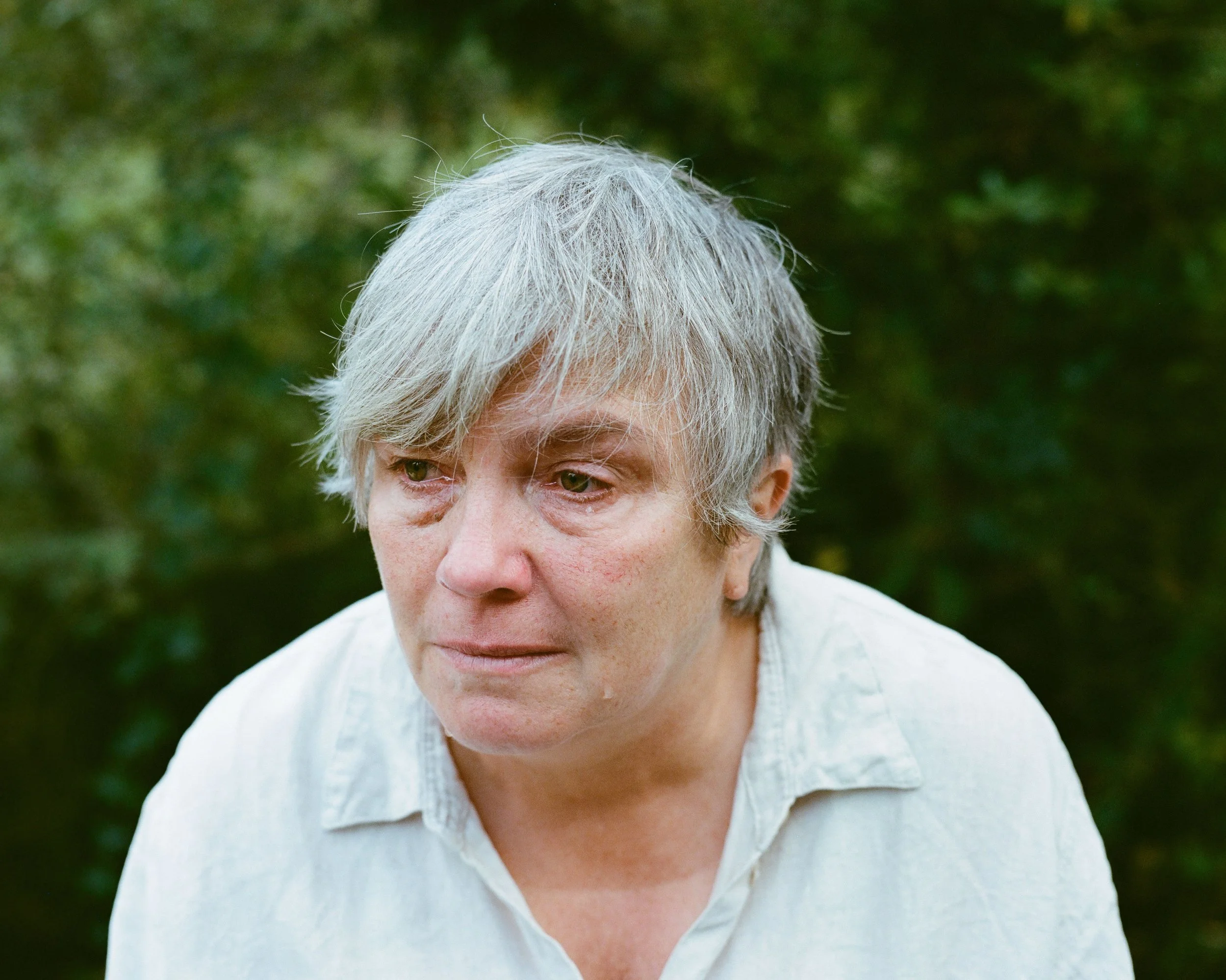 A person with gray hair, looking down with a sad or contemplative expression, outdoors with green foliage in the background.