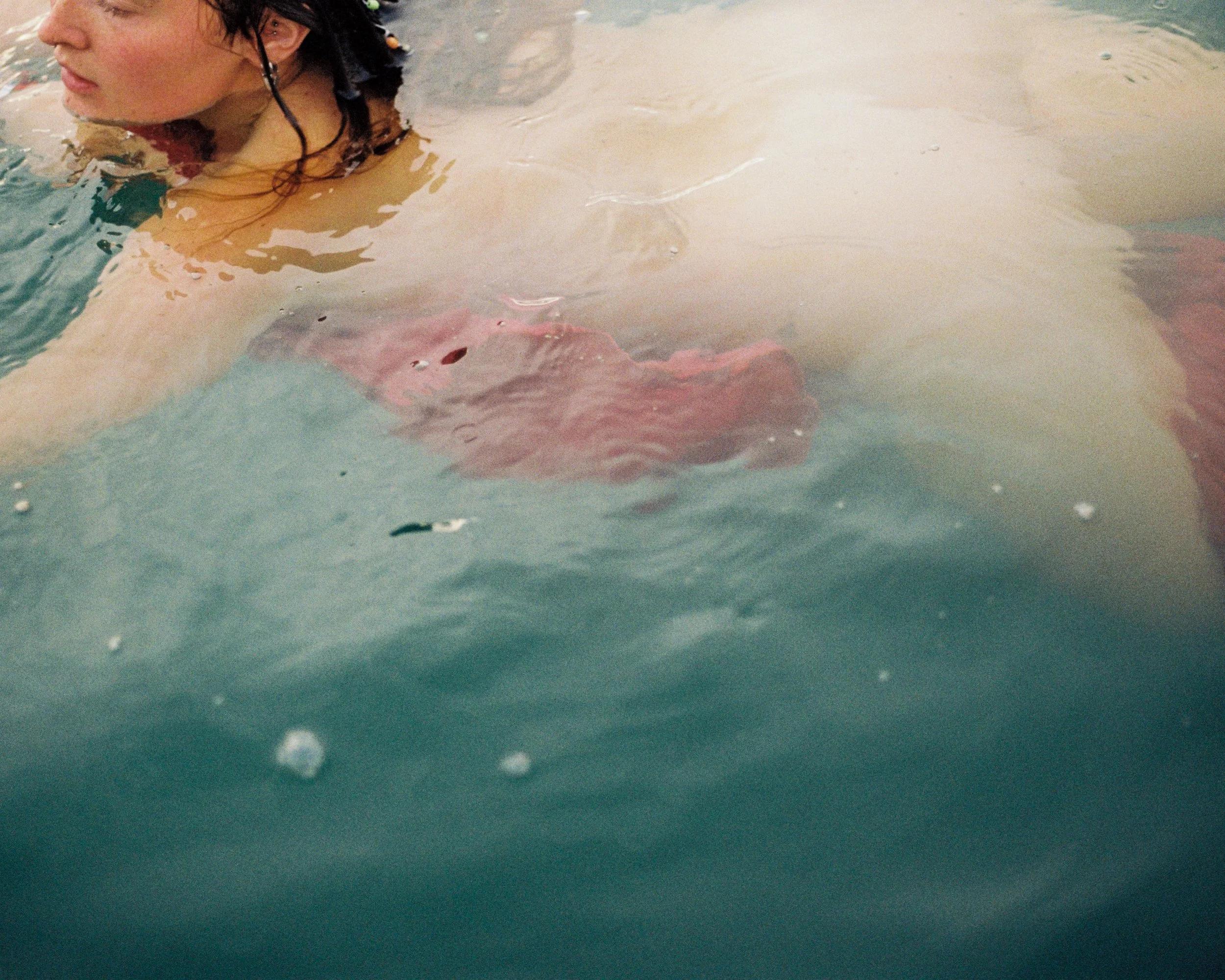 A person swimming in water, partially submerged, with their face turned to the side and dark hair wet.