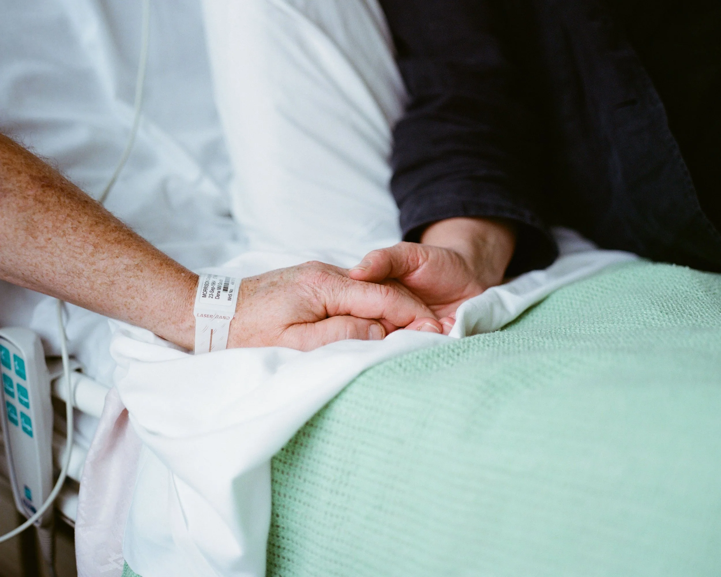 A patient and a visitor hold hands on a hospital bed, with the patient's hospital wristband visible.