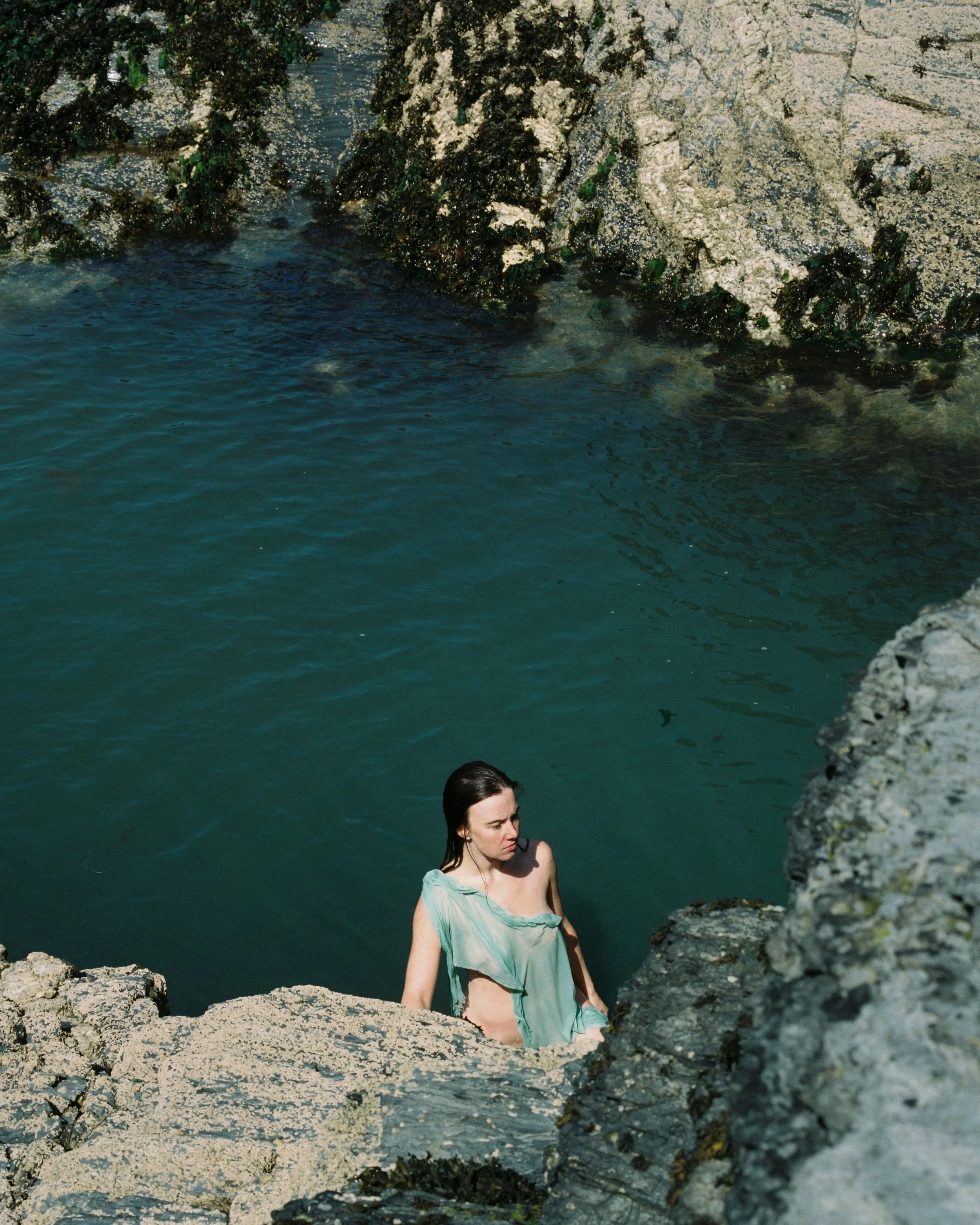 A woman with wet hair and wearing a light green dress is standing on rocks by a body of water, with rocky shoreline in the background.