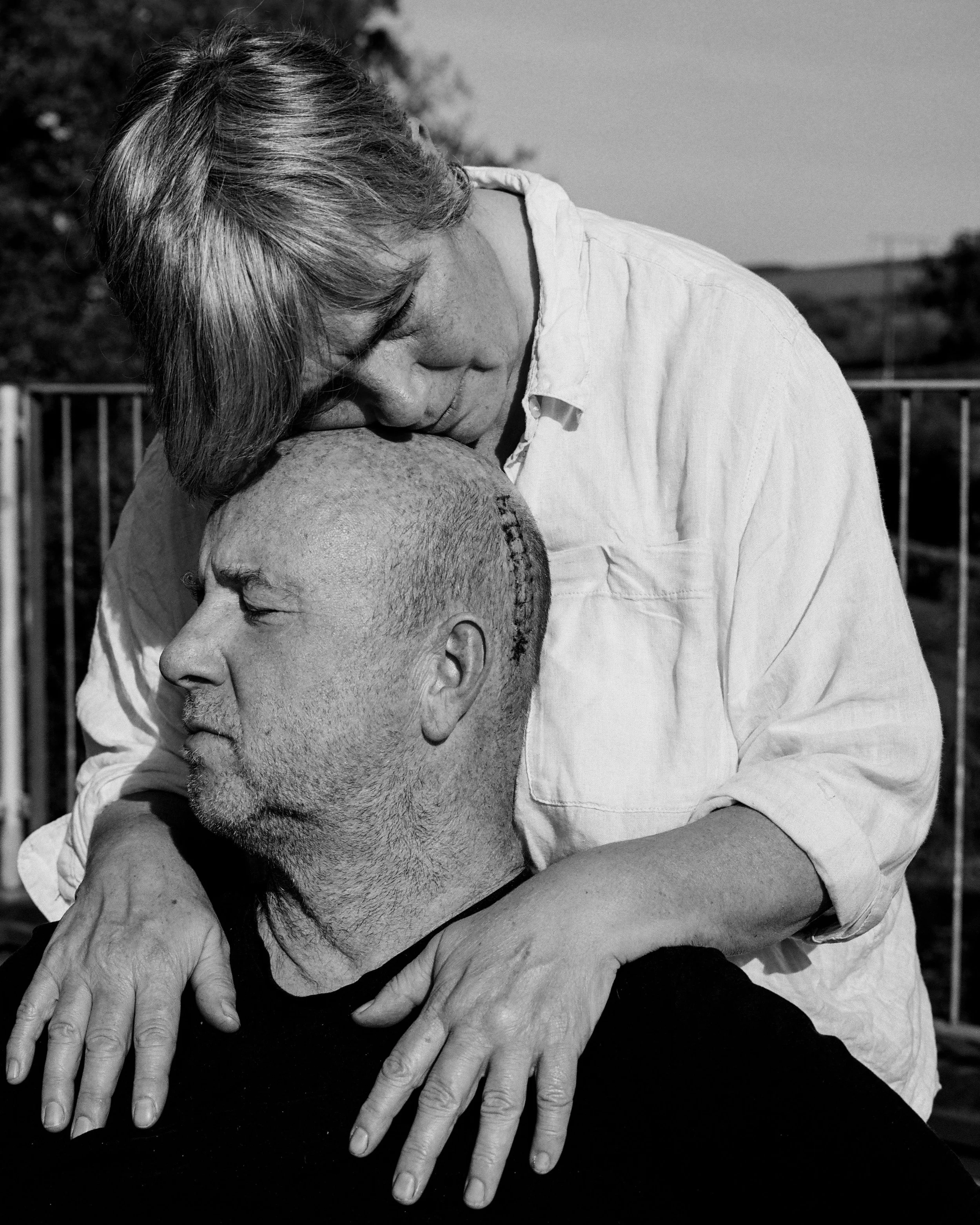 A black-and-white photo of a woman and man in an emotional embrace, with the woman resting her head on the man's head and holding his shoulders, appearing to be comforting or consoling him outdoors.