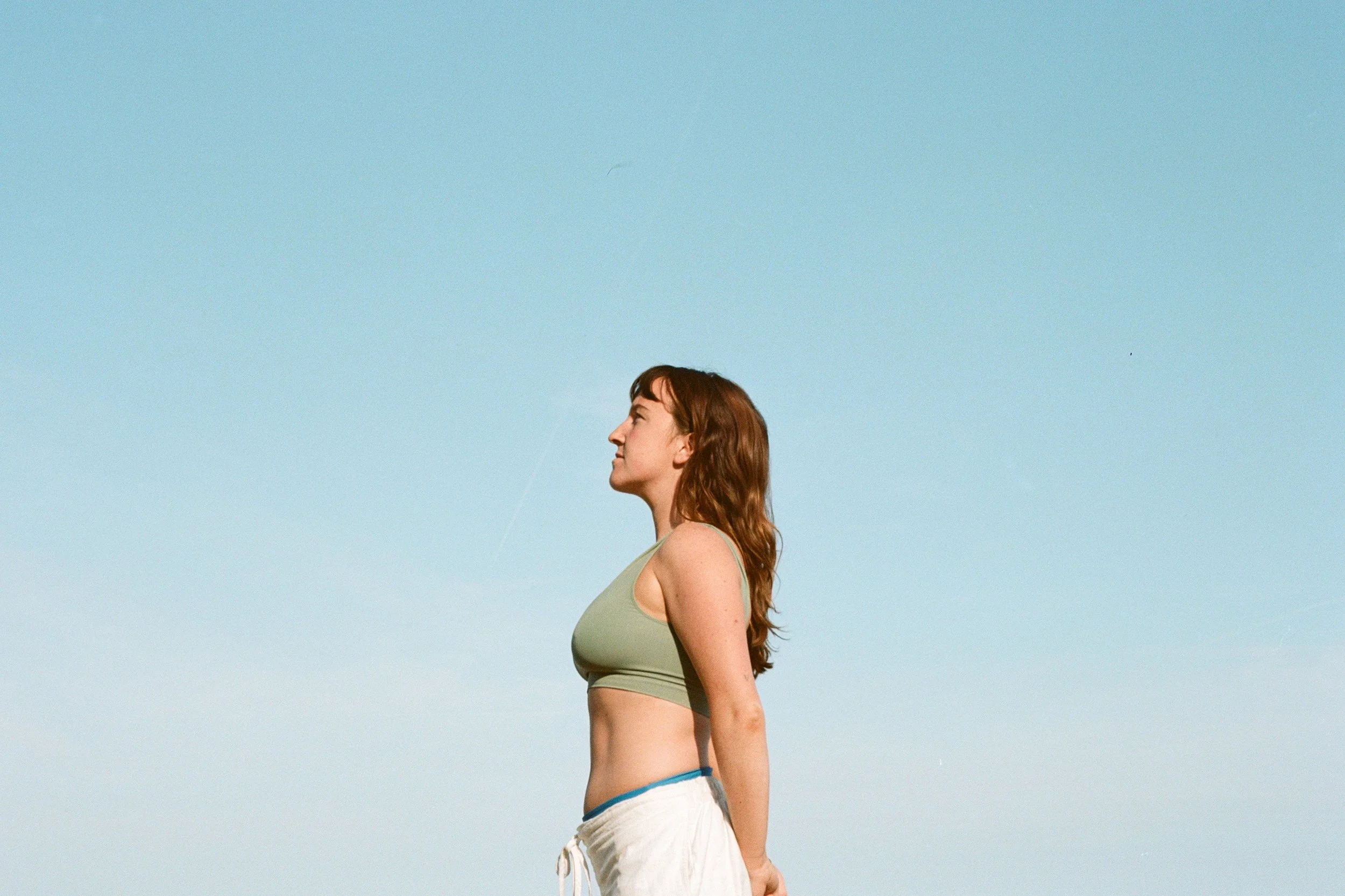 A woman with red hair standing outdoors against a blue sky, facing left with her hands behind her back.