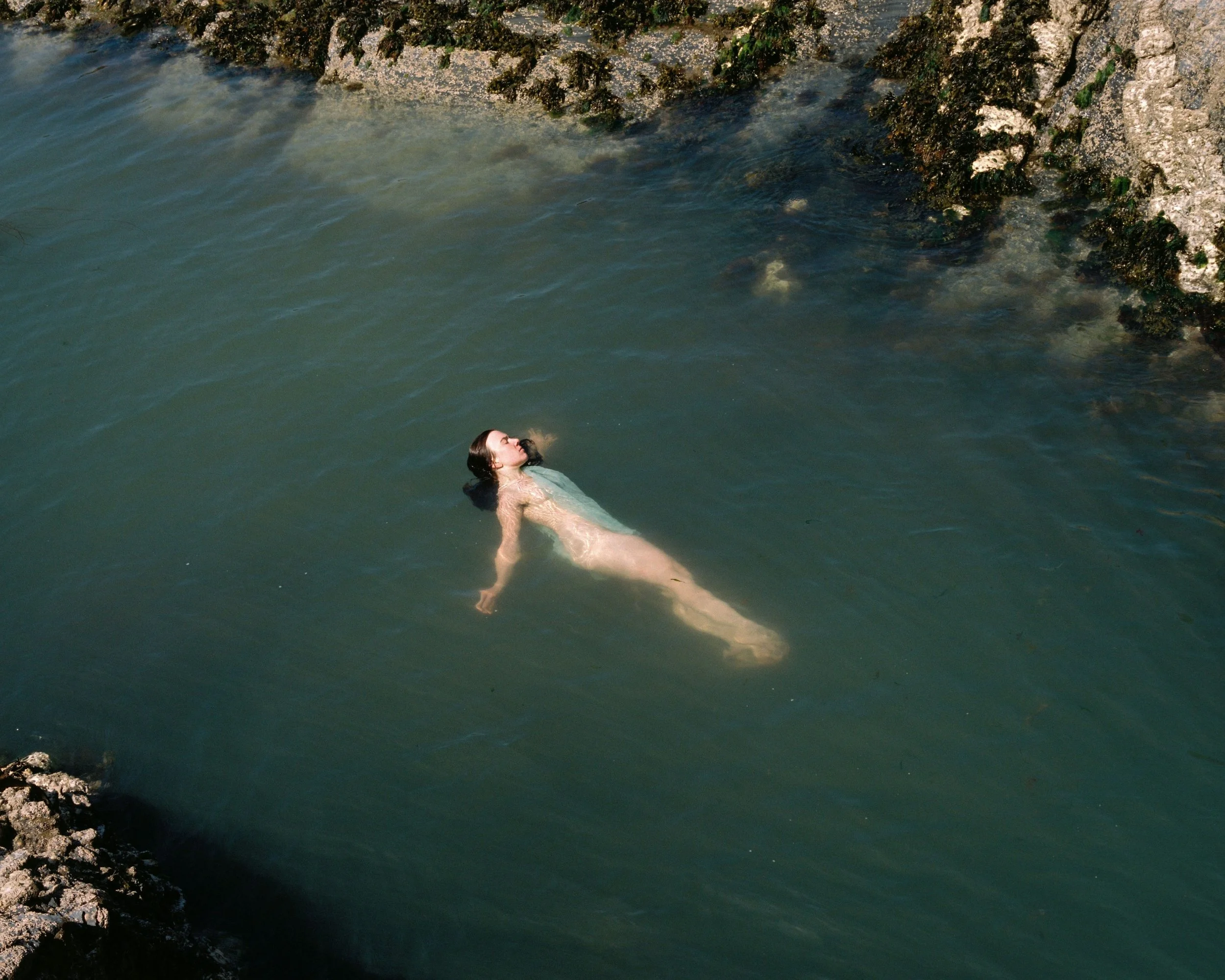 A nude woman floating on her back in a body of water near rocky shoreline.