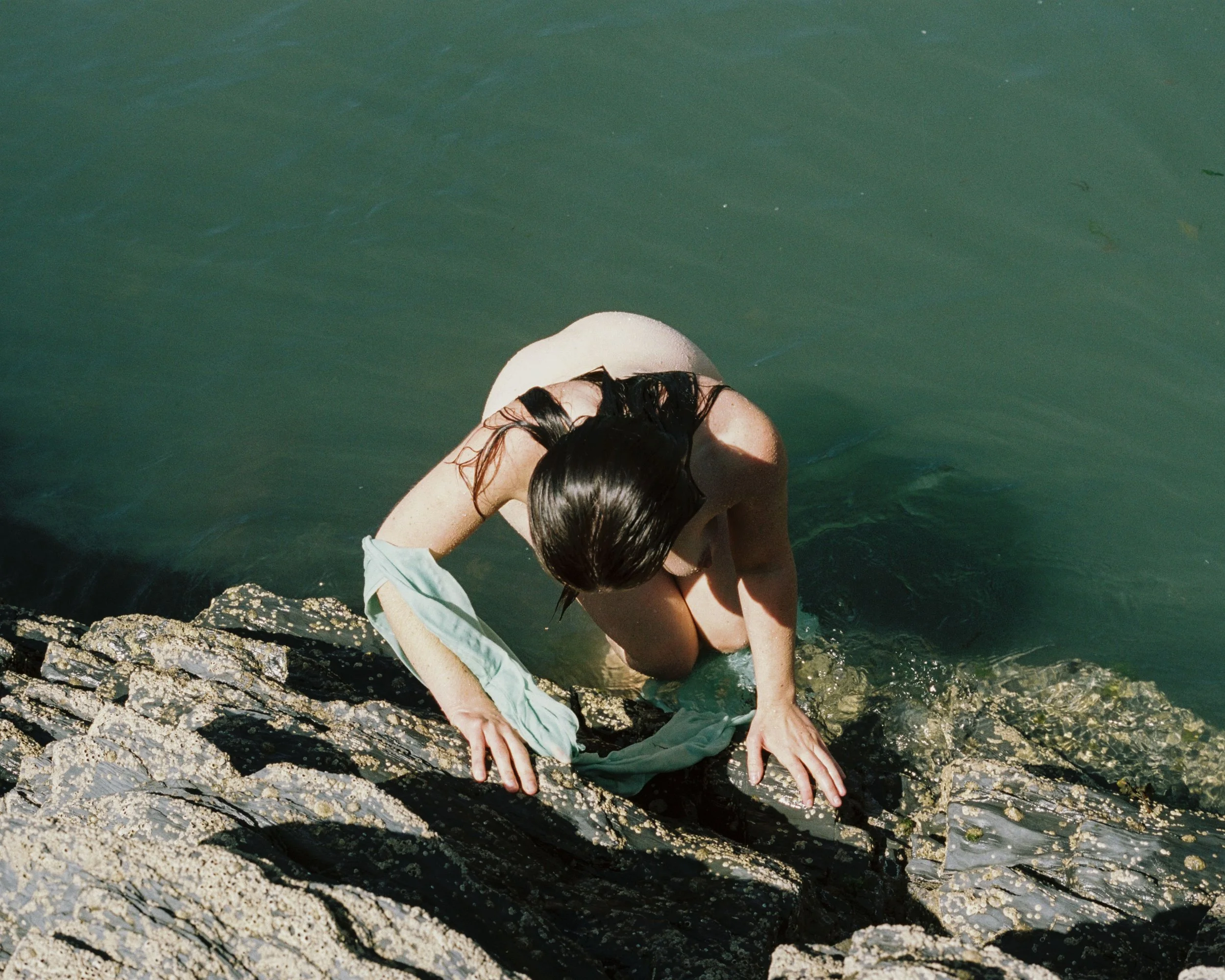 A person with long dark hair, wearing a light-colored swimsuit, climbing out of a greenish body of water onto a rocky shore, holding a towel.