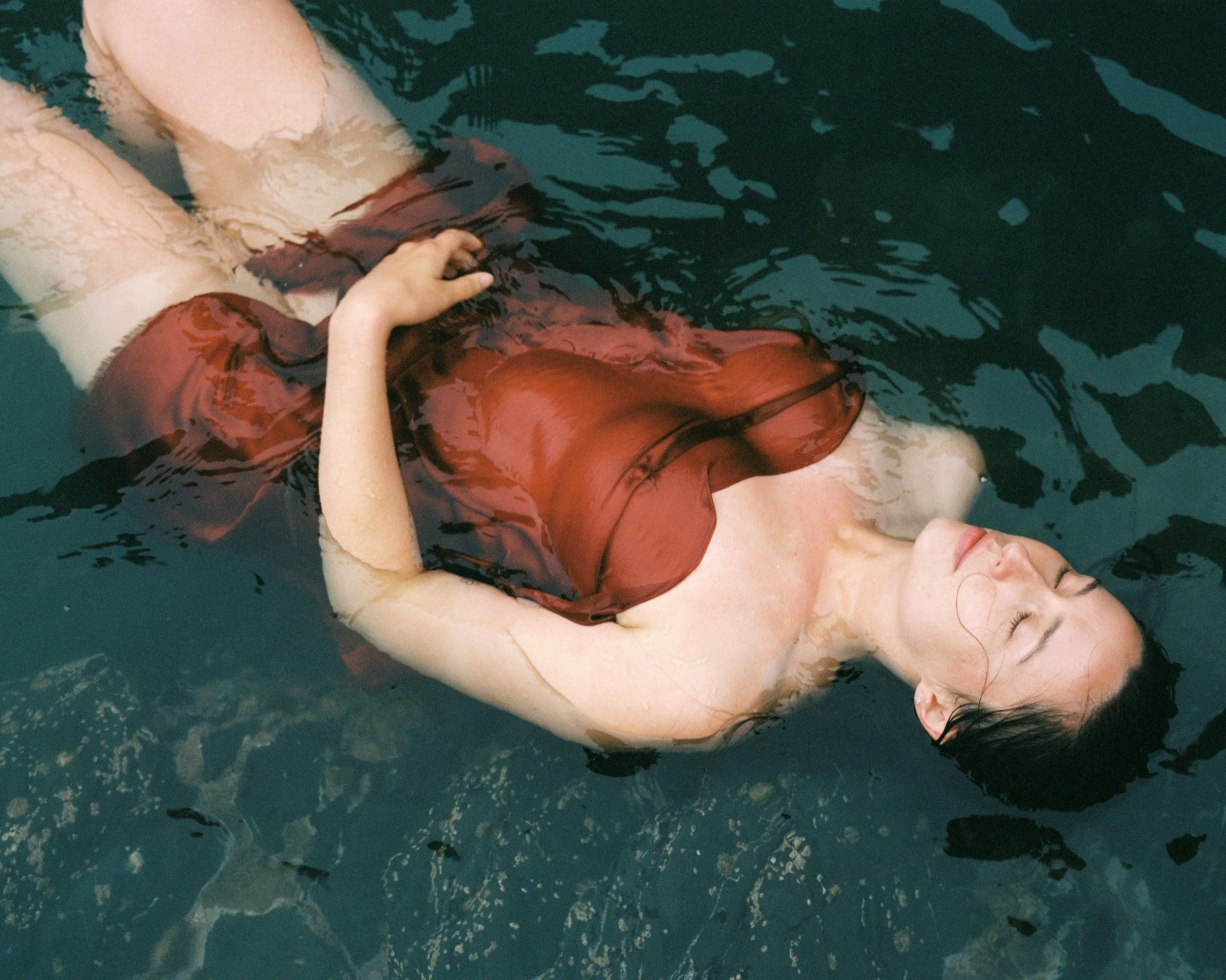 A woman floating on her back in a dark swimming pool, wearing a sheer red top and light-colored bottoms, with her eyes closed and arms relaxed.