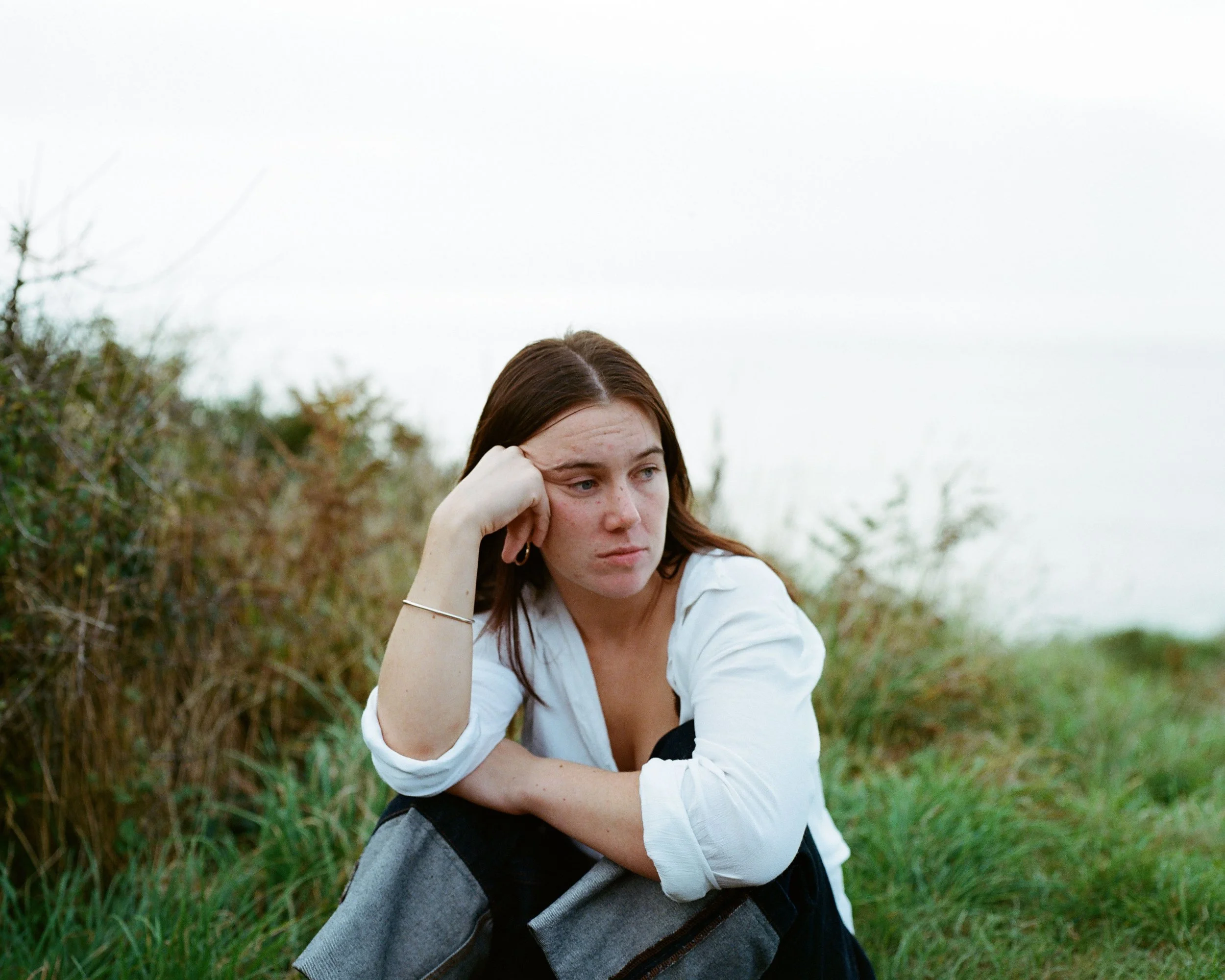 A woman sitting outdoors among grass and shrubs with a somber expression, resting her head on her hand.