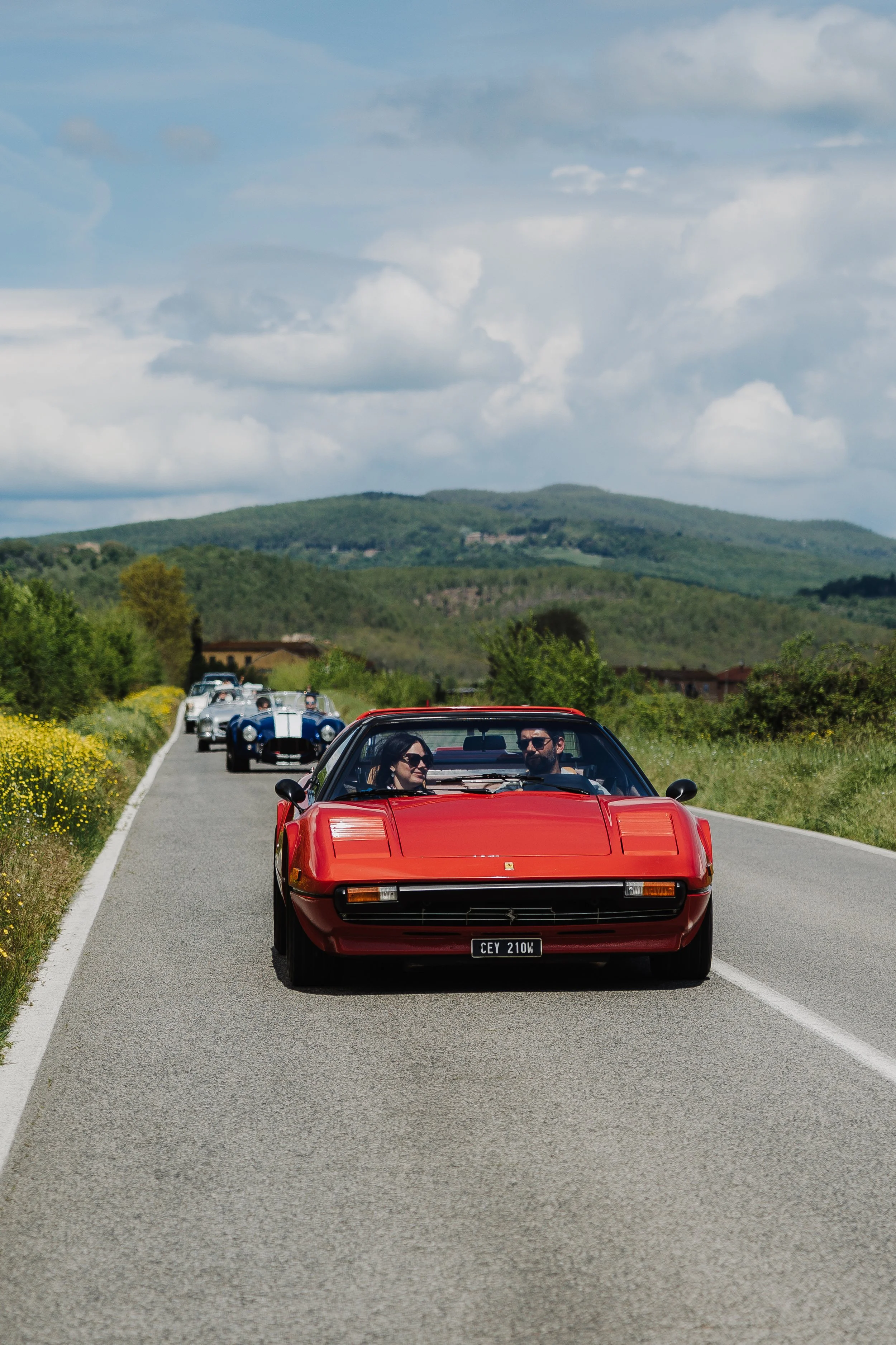 A line of vintage sports cars driving on a scenic countryside road with green hills and partly cloudy sky in the background, led by a red Ferrari with two passengers.