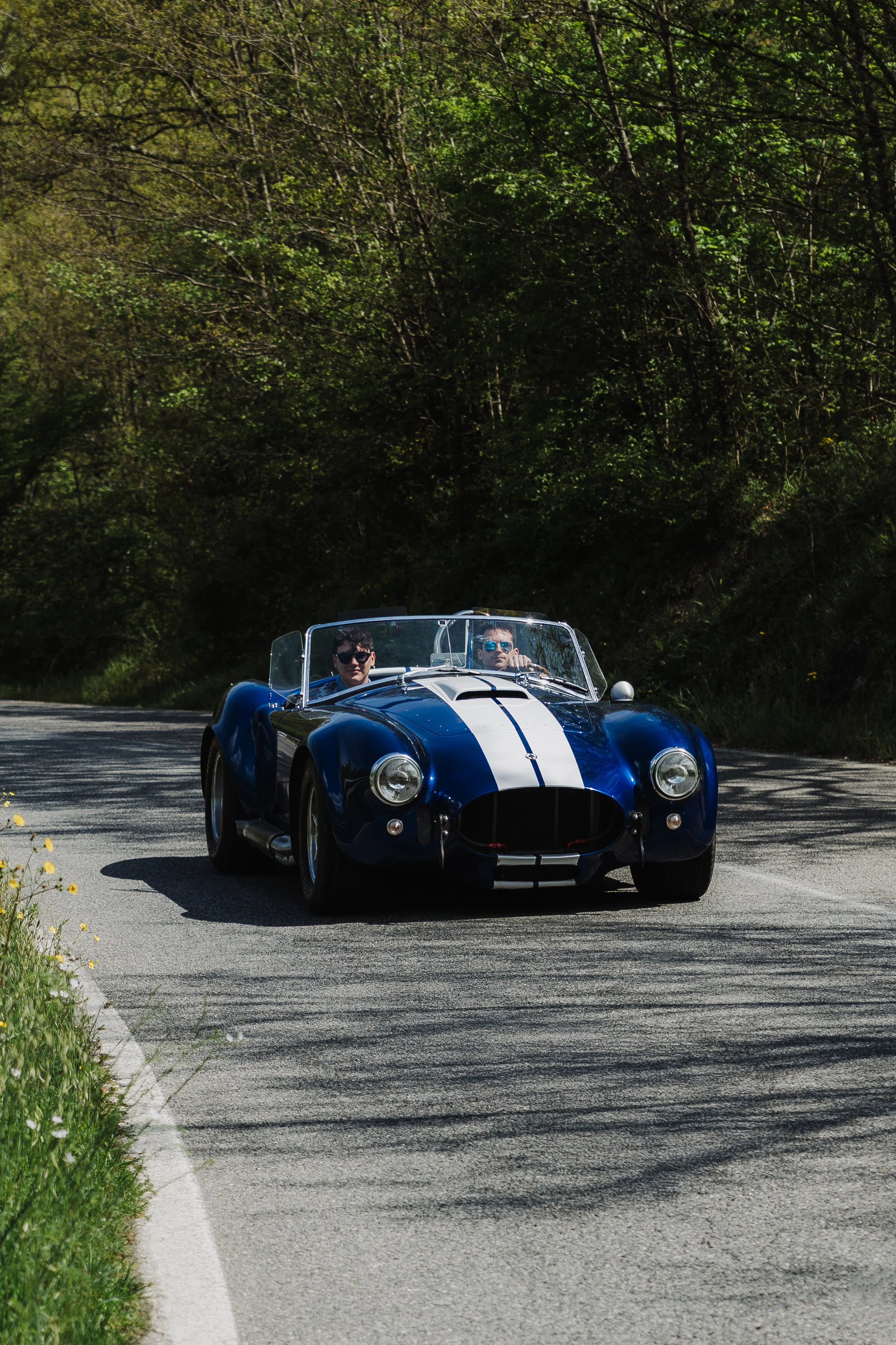 A blue vintage sports car with white racing stripes, driven by two men wearing sunglasses, on a winding rural road surrounded by green trees.