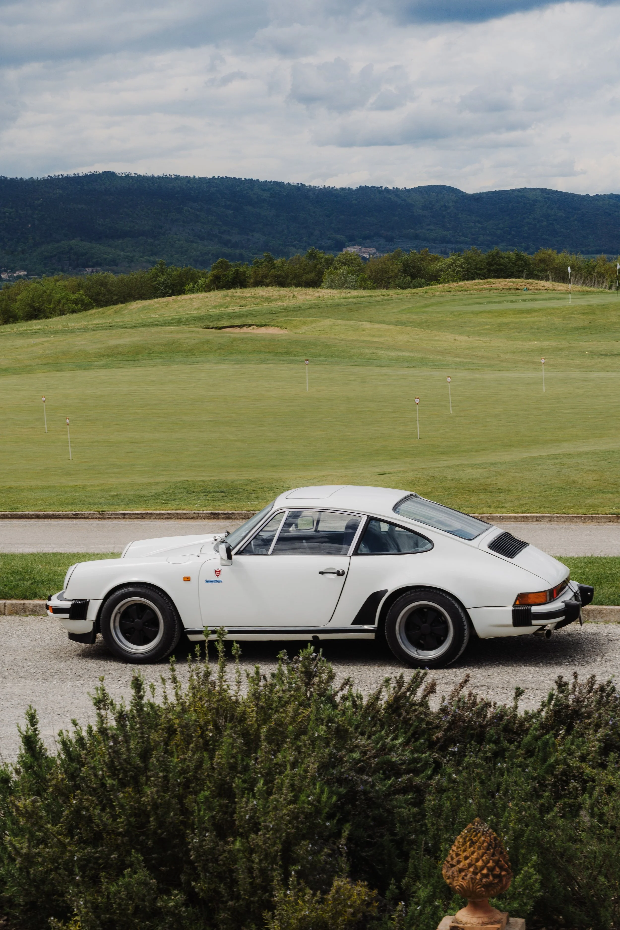 A white vintage Porsche sports car parked on a driveway with a golf course and green hills in the background under a cloudy sky.