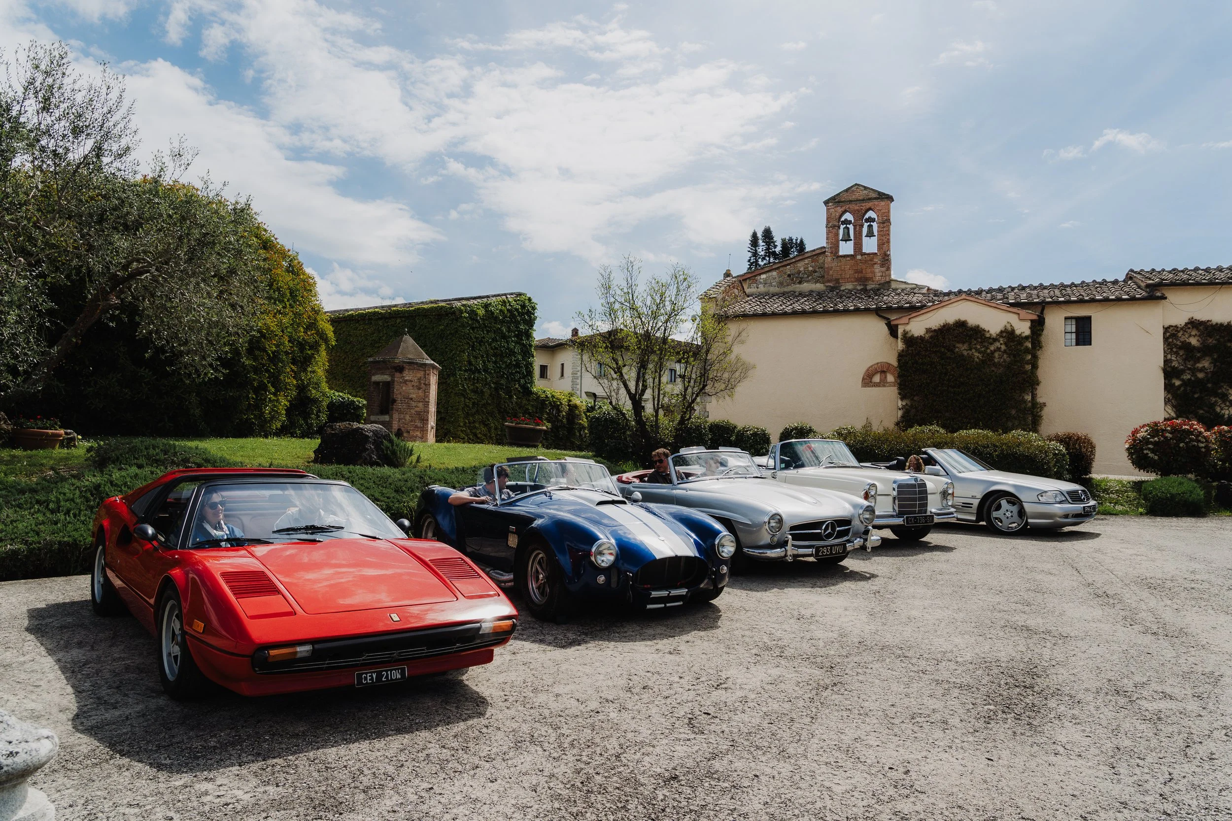 Five classic cars parked in a row in front of an Italian-style building with a bell tower, with people inside the cars, and trees and greenery in the background.