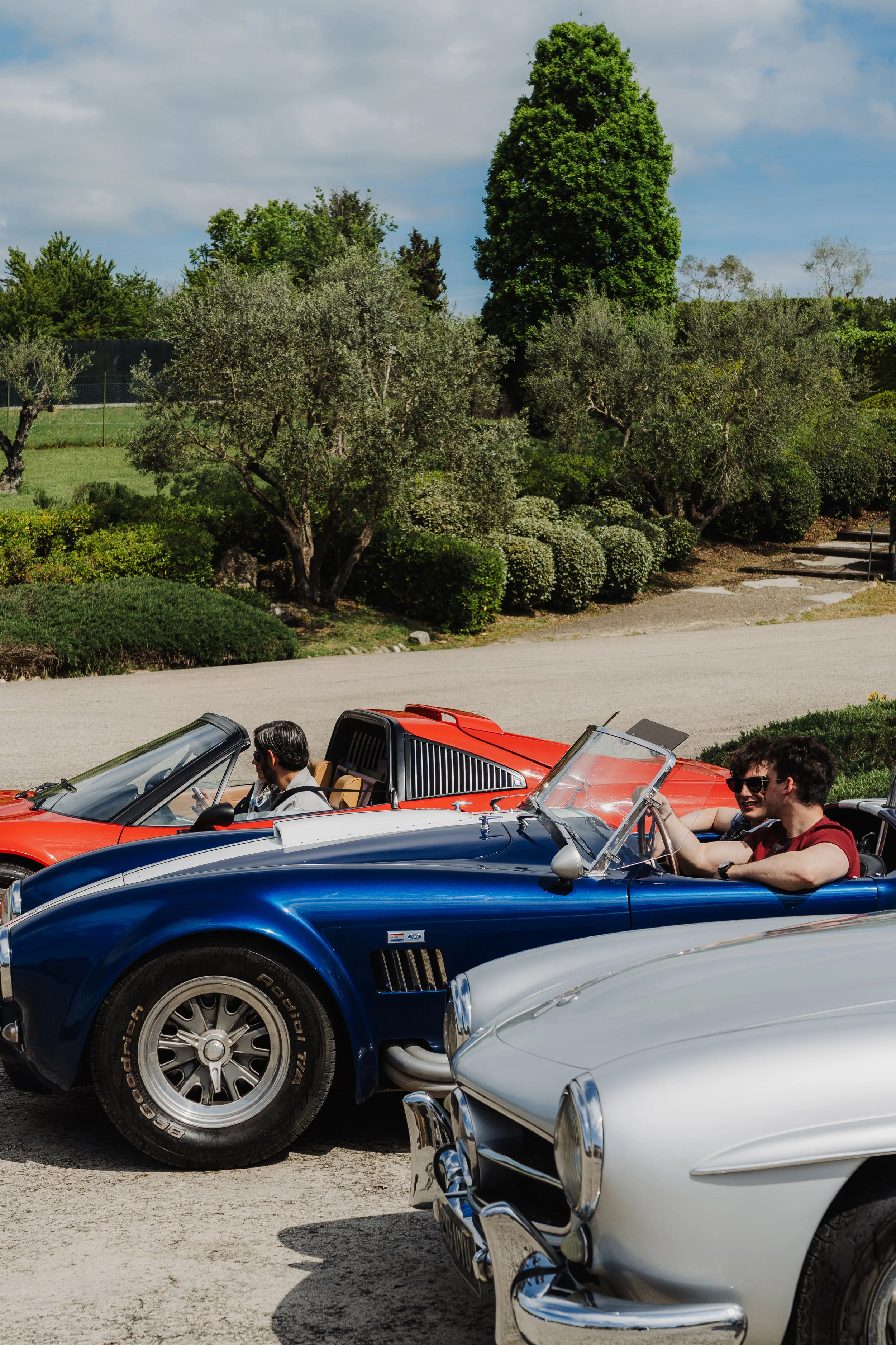 Three vintage sports cars in blue, red, and white parked outdoors with drivers and passengers enjoying the sunny day, trees and greenery in the background.