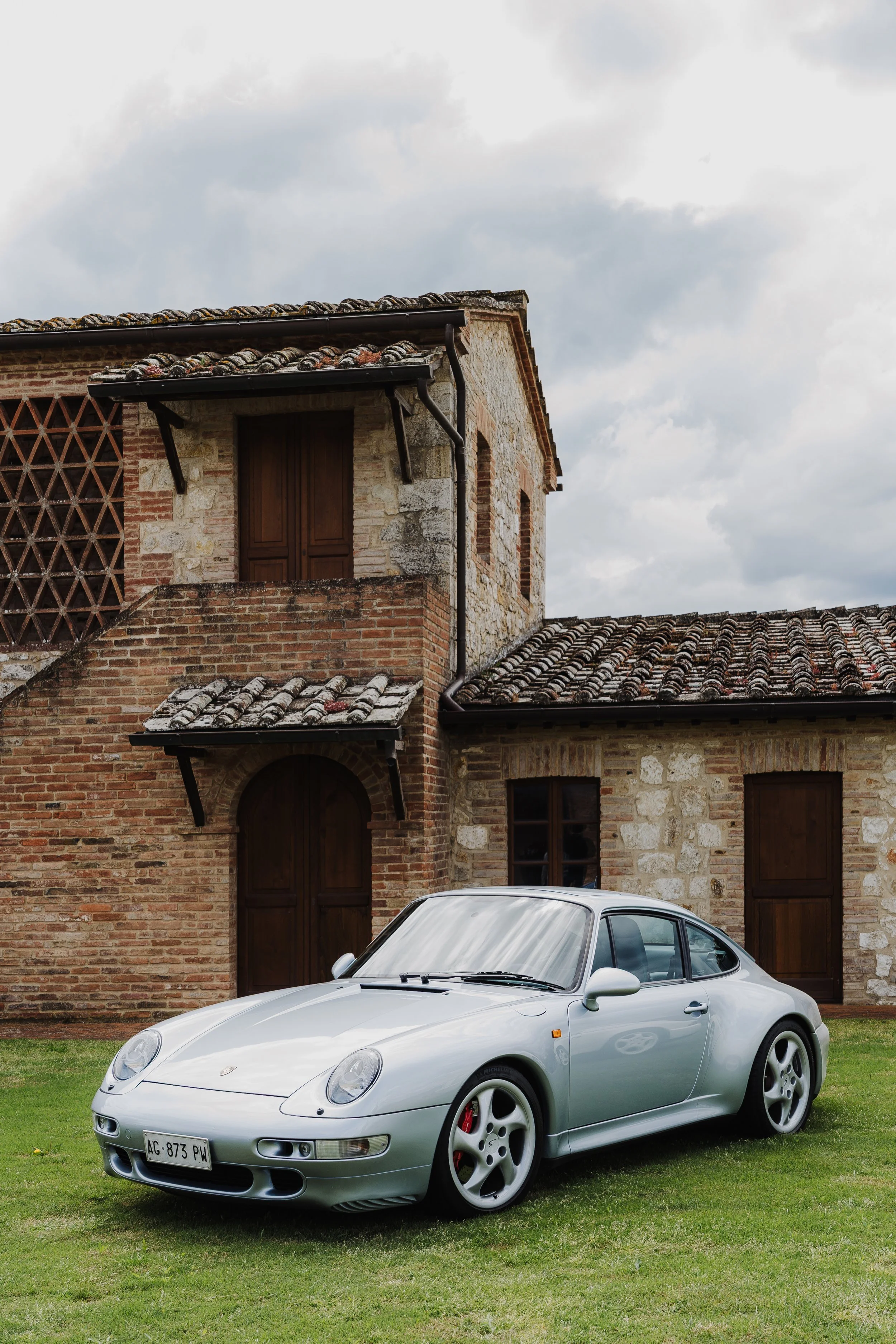 A silver Porsche sports car parked on a lawn in front of a rustic brick and stone house with wooden shutters.
