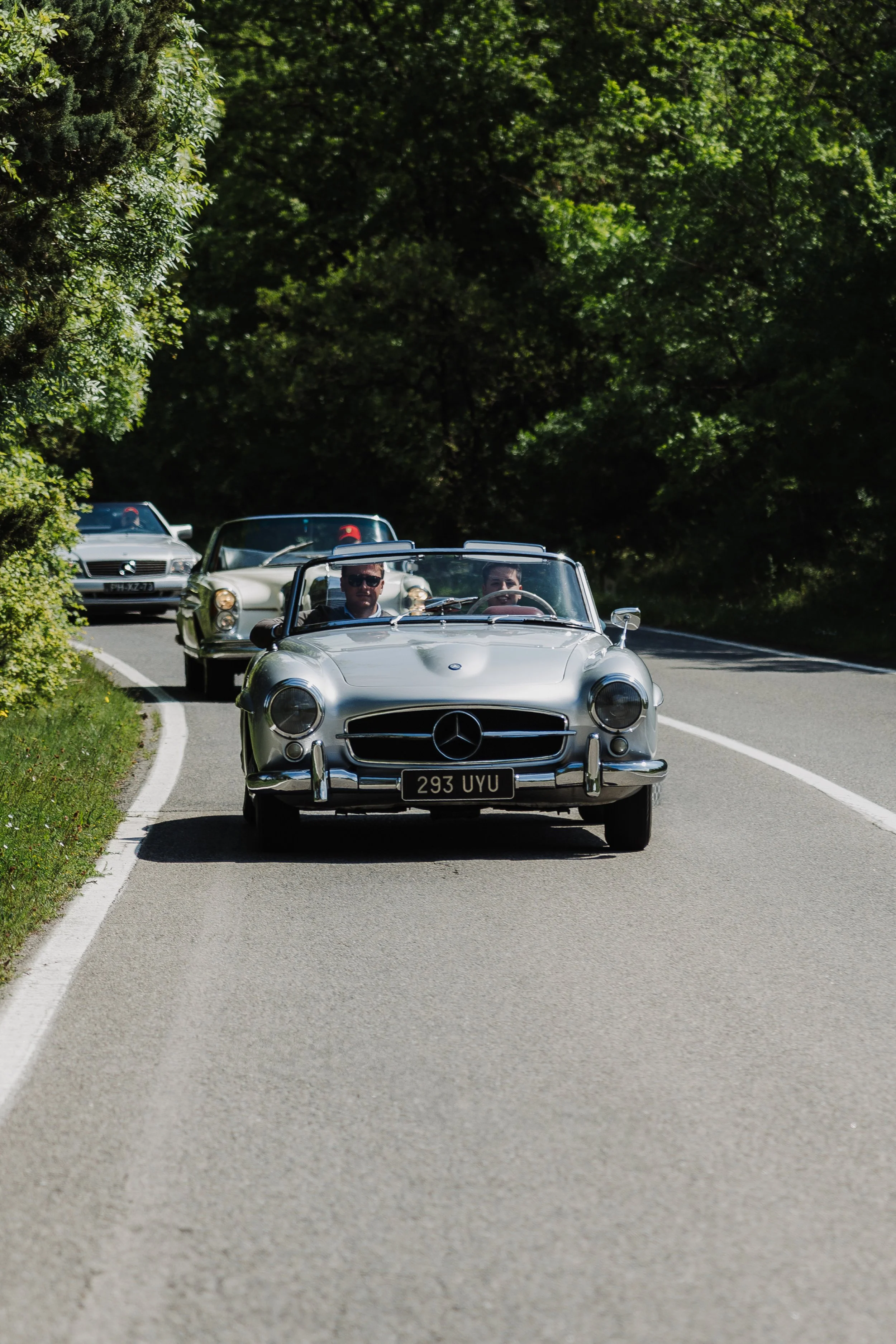 Three vintage Mercedes-Benz cars driving on a winding rural road with green trees on both sides, with the front car being silver and the other two darker-colored, two people seen in the front car, one wearing sunglasses.