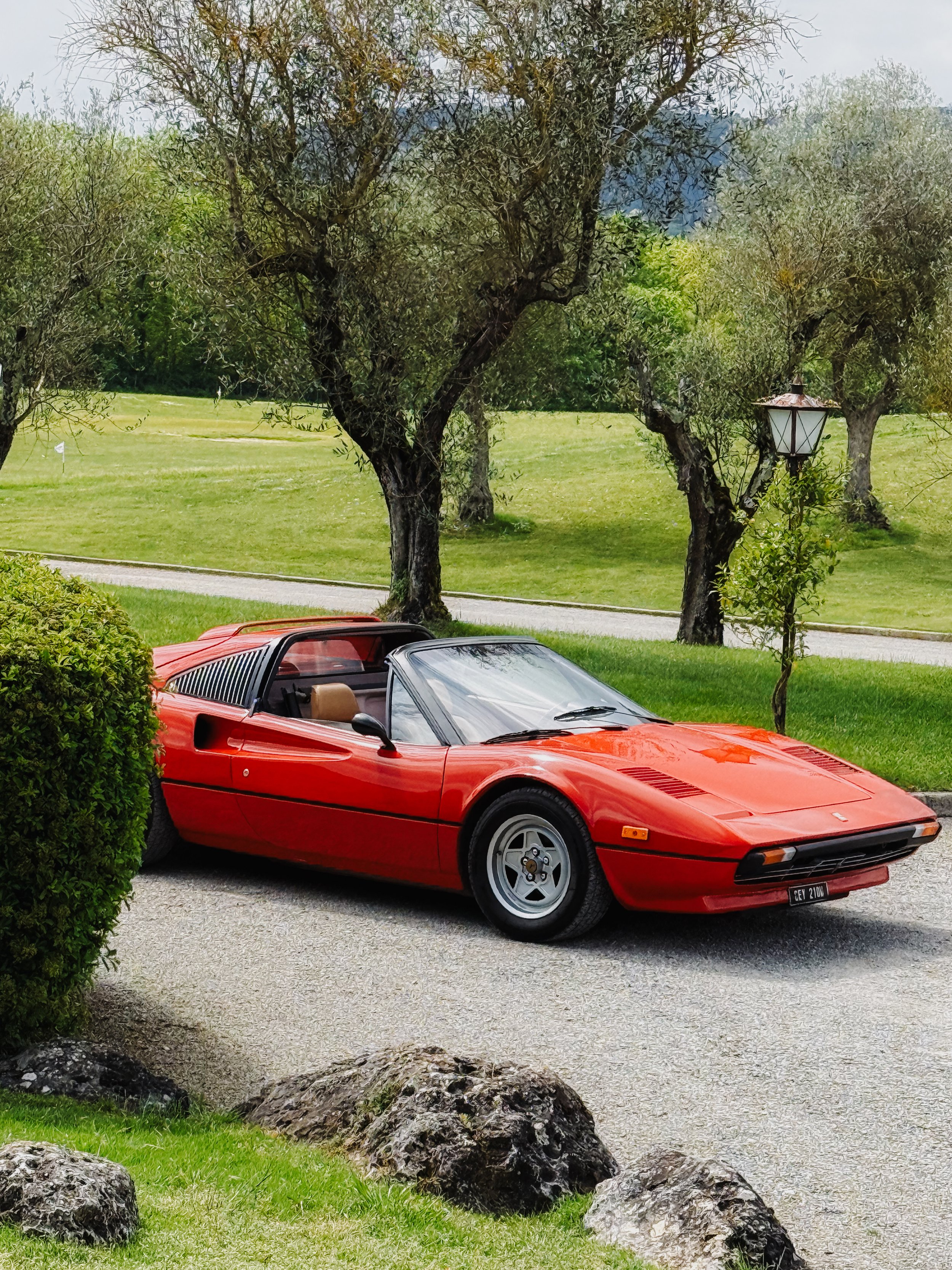 A red vintage Ferrari sports car parked on a gravel driveway next to a green lawn with trees and a lamppost in the background.
