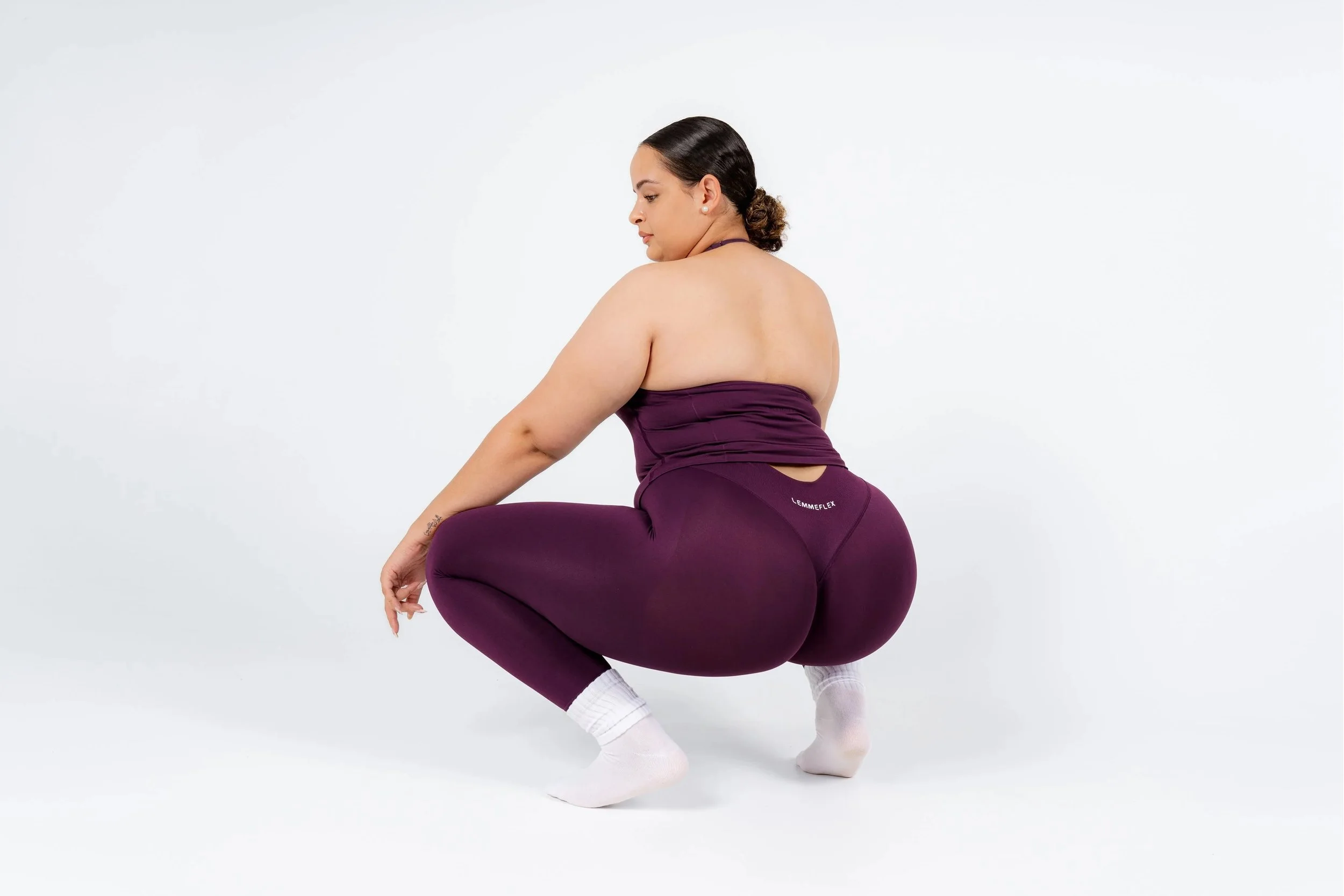 A woman in purple workout attire, wearing white socks, is squatting with her back facing the camera in a white studio setting.
