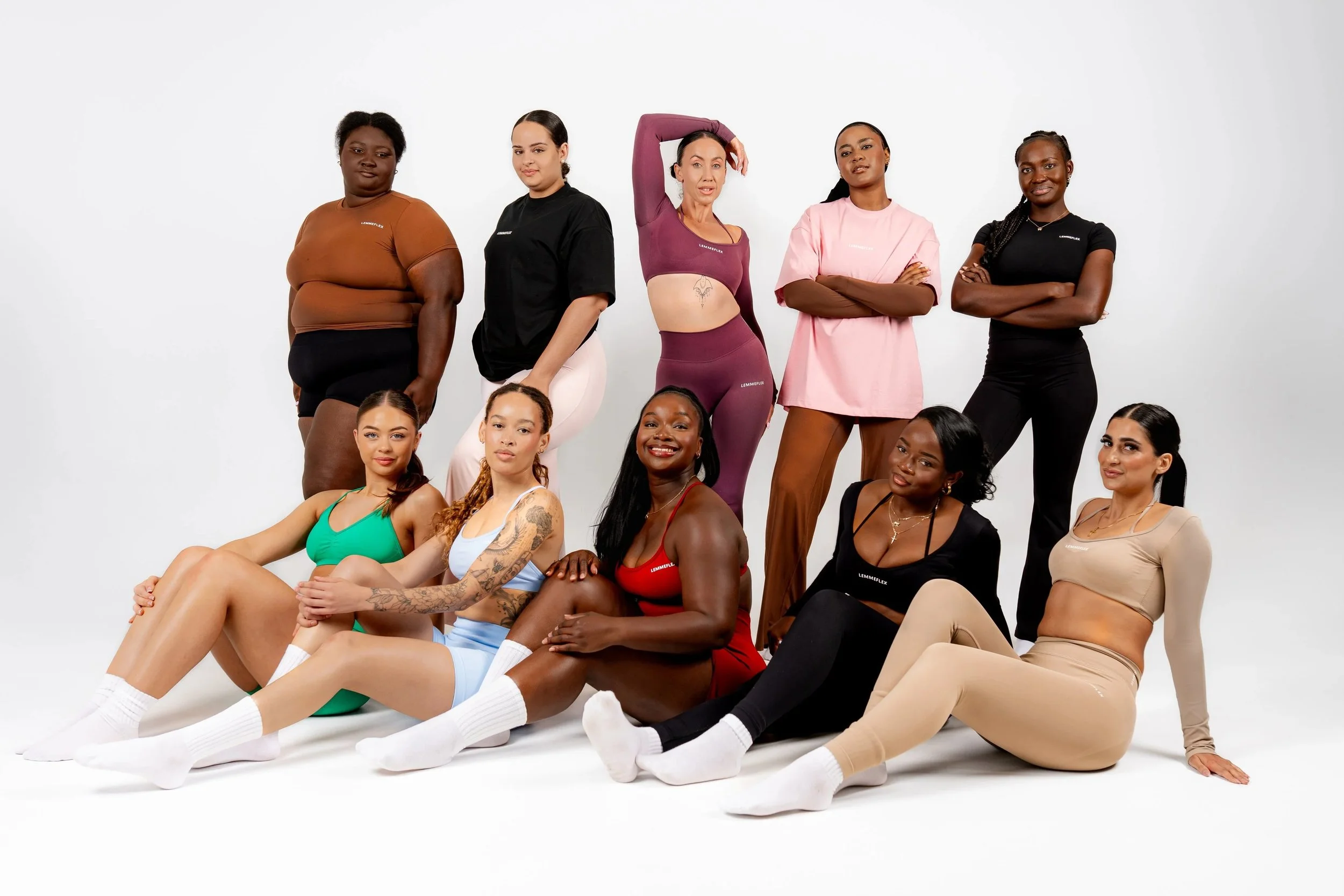 A diverse group of women posing together against a white background, some sitting and some standing, wearing casual and athletic clothing.