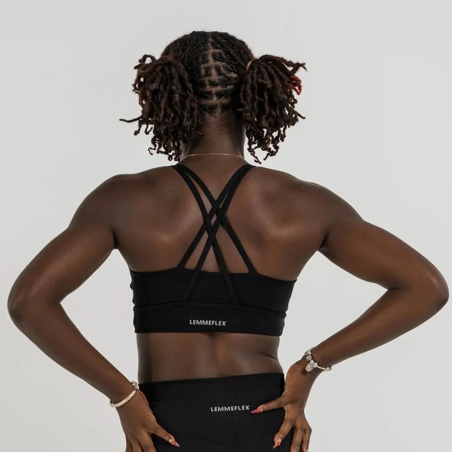 Back view of a woman with dark complexion and dreadlocks wearing a black sports bra with the word LEMMEFLEX printed on the band.