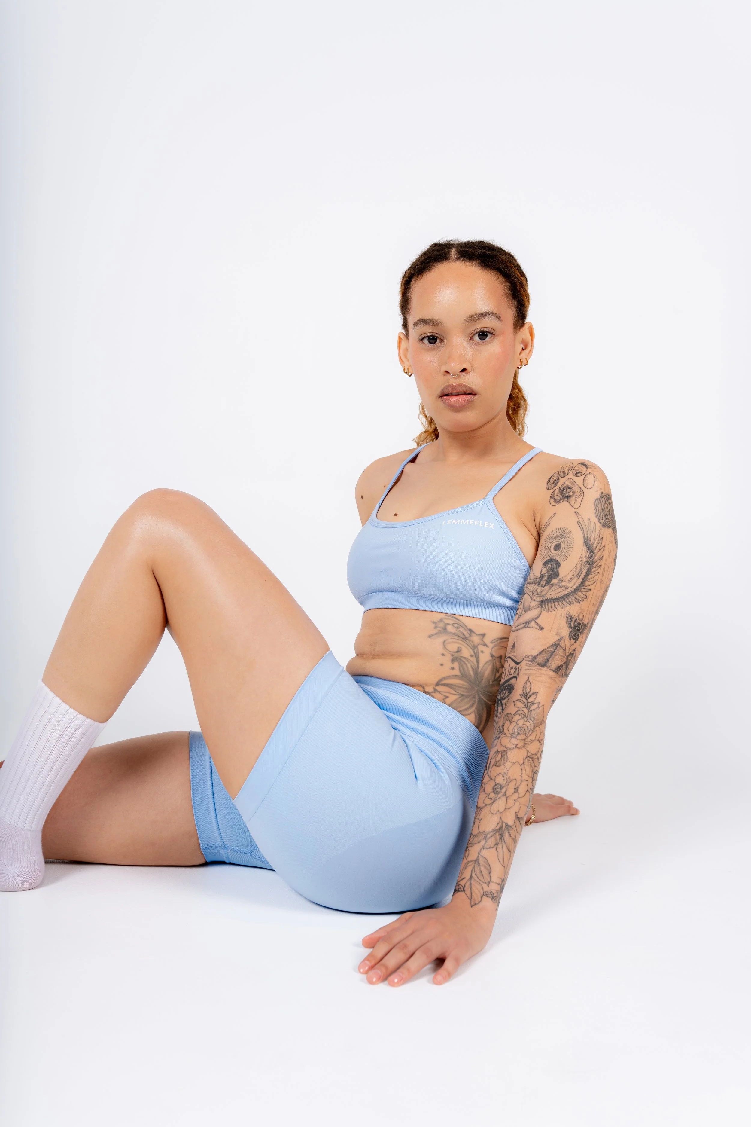 Young woman with tattoos sitting on the floor with one knee bent, wearing a light blue sports bra and matching shorts, against a plain white background.