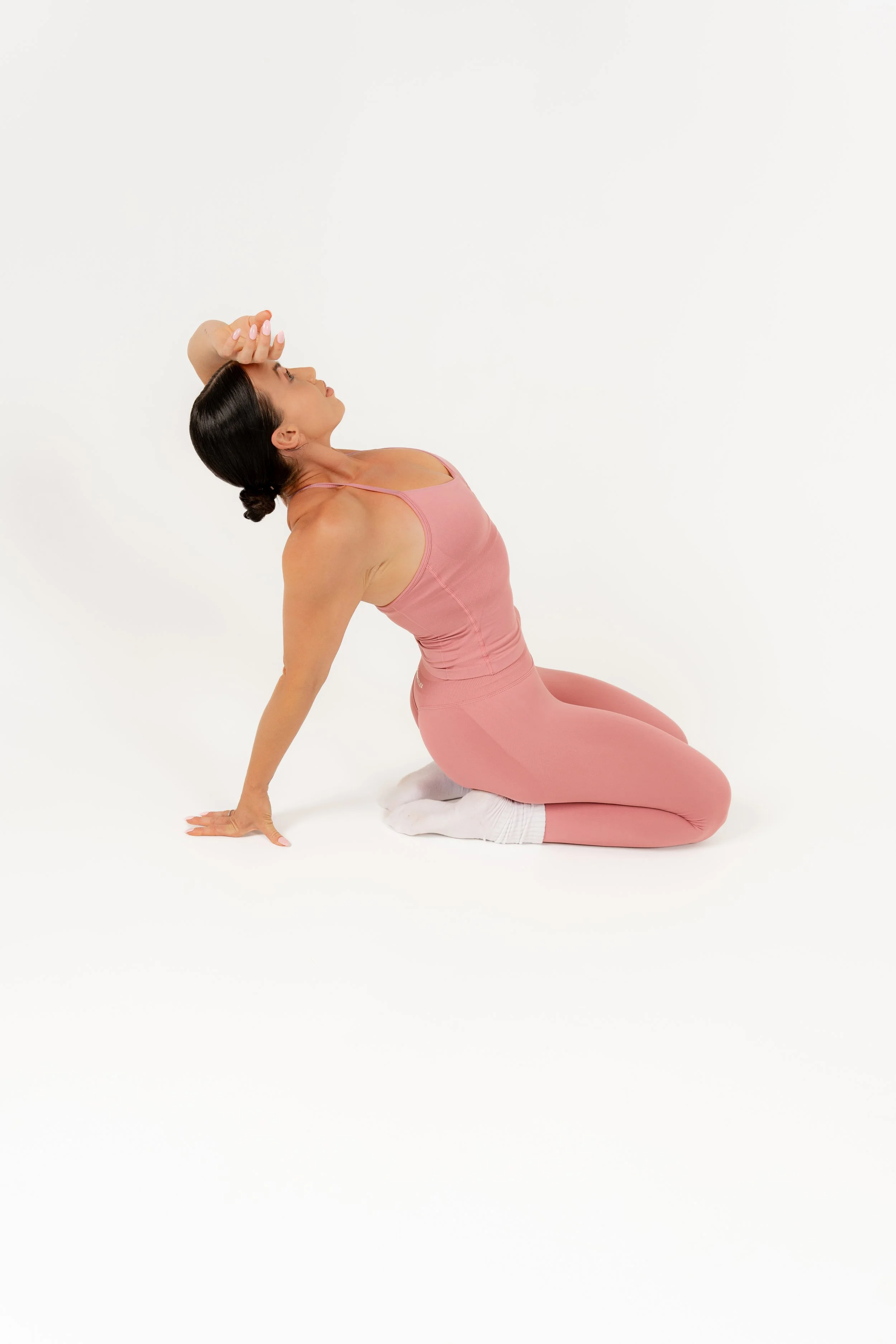 A woman performing a yoga pose on a white background, kneeling with her right hand on the floor, left hand on her forehead, looking up, dressed in a pink sports bra and matching leggings.