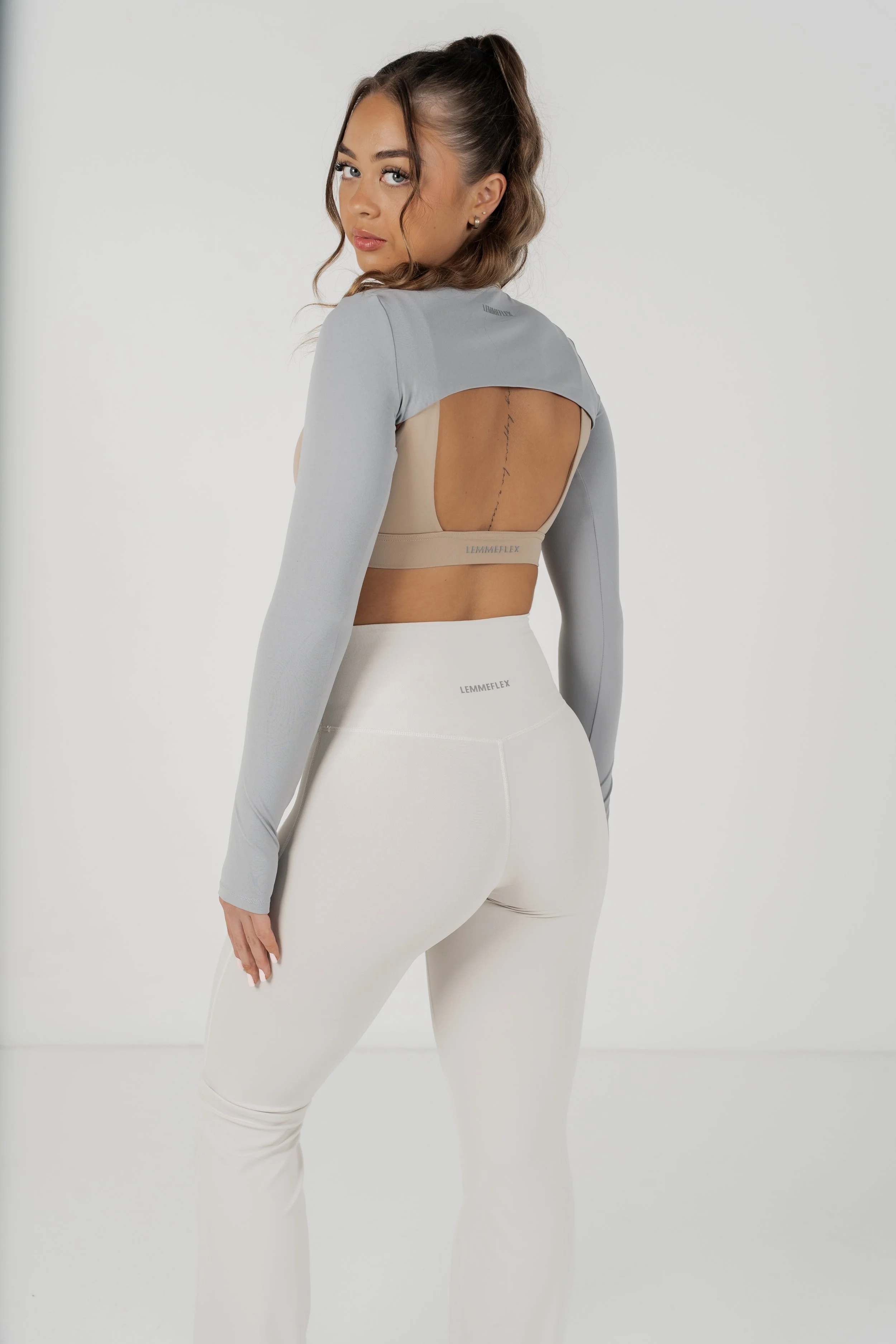 A woman with wavy brown hair tied in a ponytail, wearing a light gray sports top with an open back and matching high-waisted leggings, standing against a plain white background.