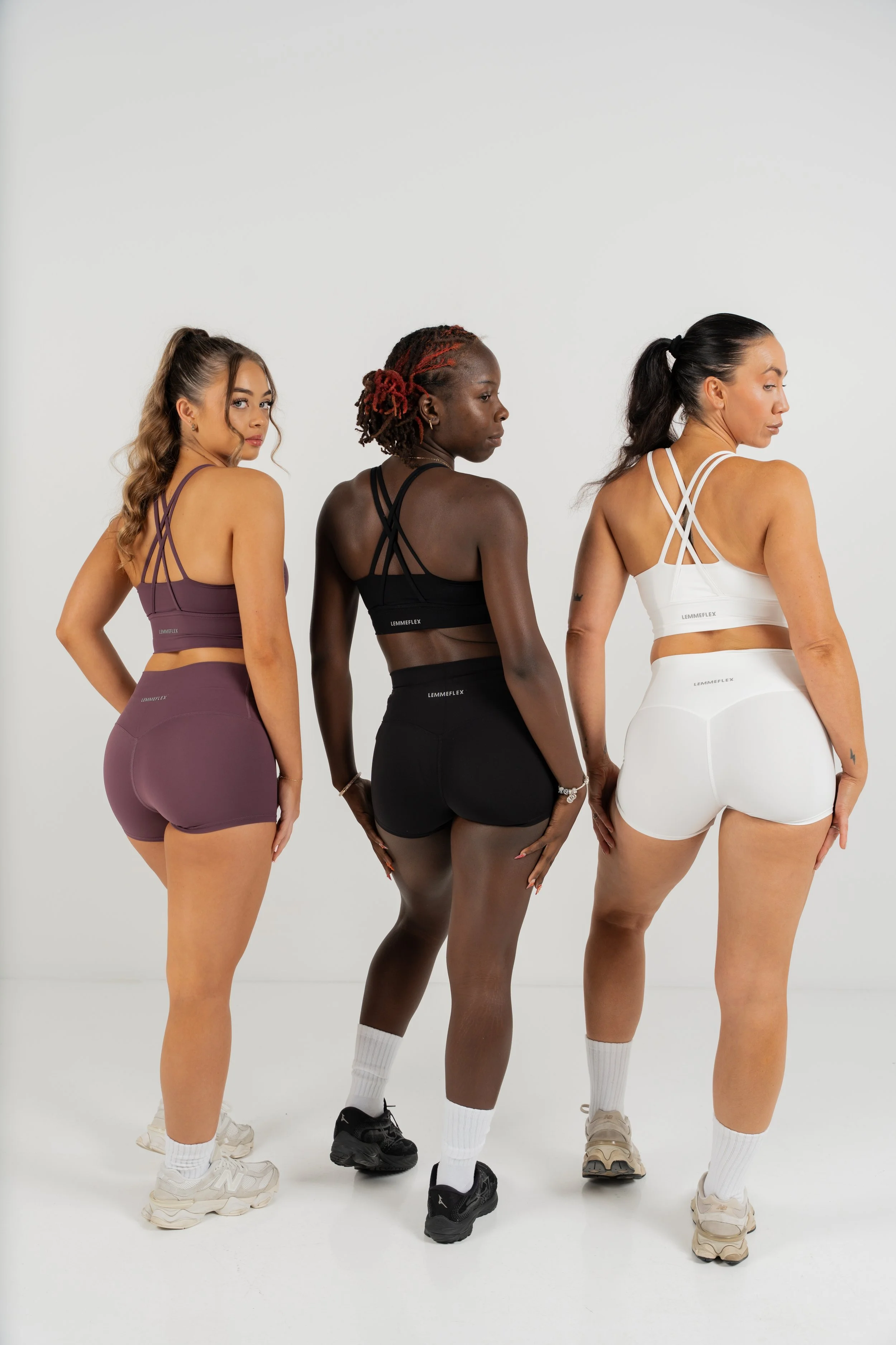 Three women standing in a row, viewed from behind, wearing athletic workout clothes in purple, black, and white, with different hairstyles and sneakers, against a plain white background.