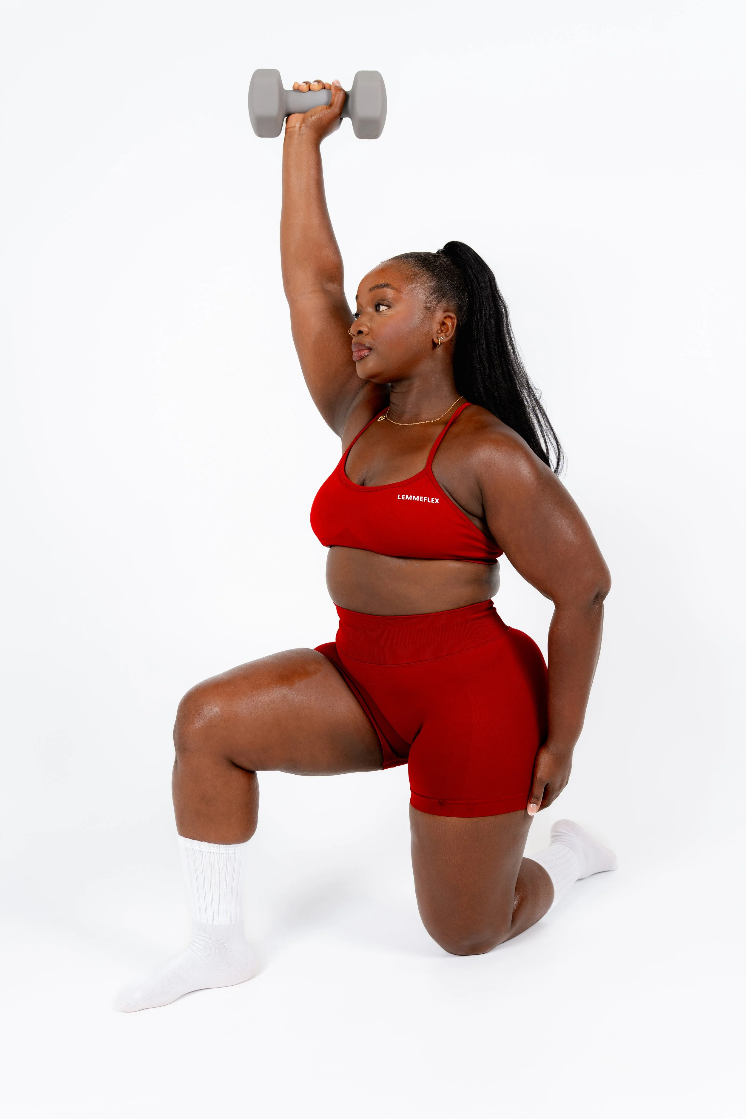A woman with dark skin in red workout clothing is kneeling on one knee with her left foot flat on the ground, holding a gray dumbbell in her raised right hand above her head, against a white background.