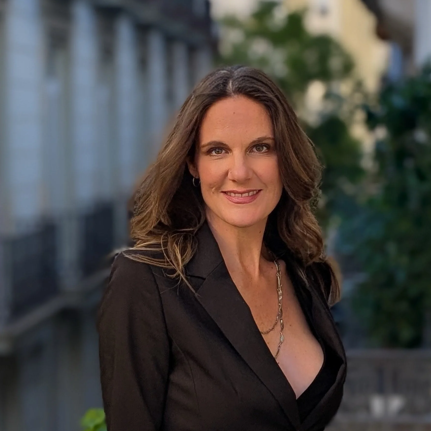 Professional headshot of a smiling woman with long brown hair wearing a black blazer and layered necklace