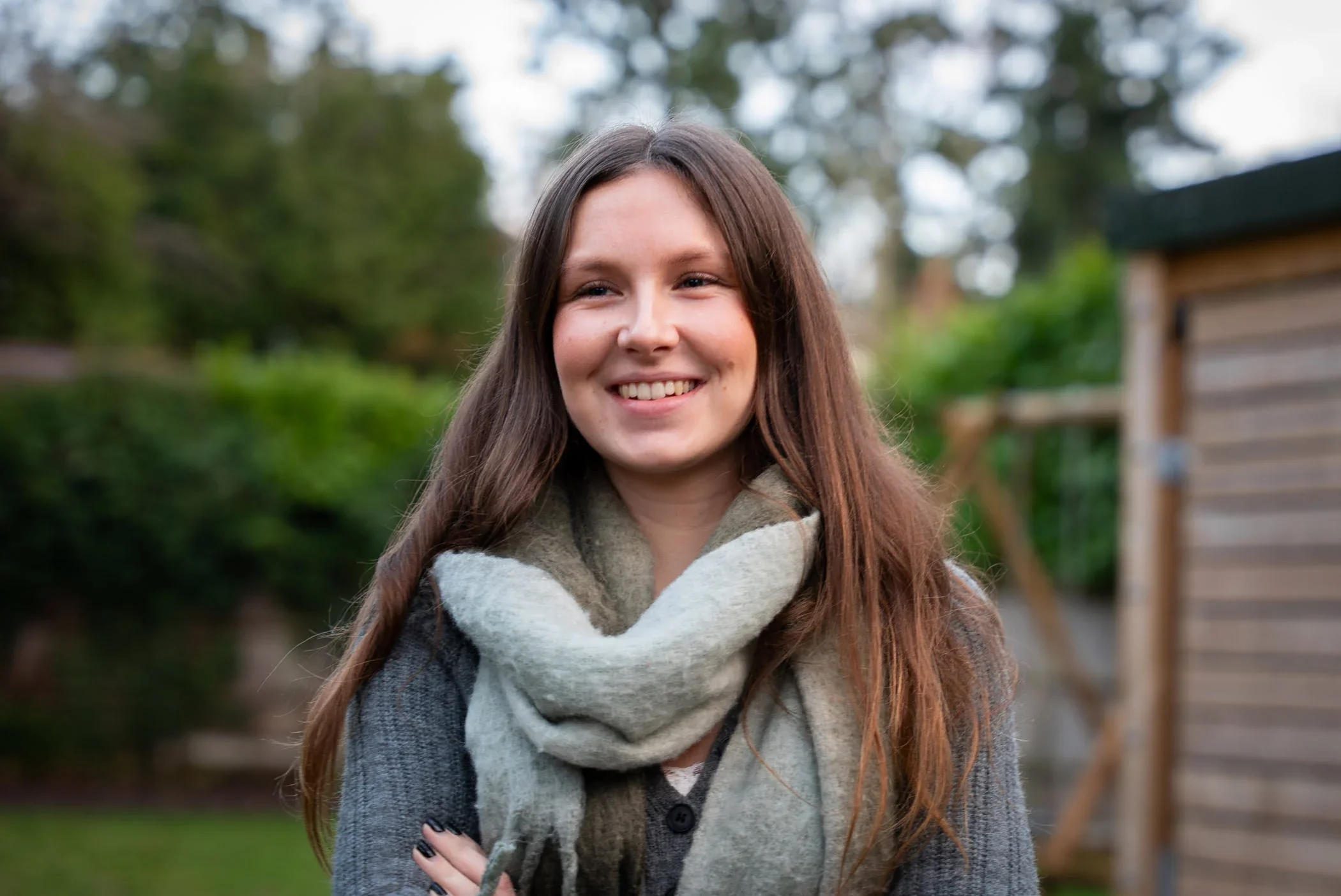 A young woman with long brown hair smiling outdoors, wearing a gray coat and a large plaid scarf, in a garden or backyard setting.