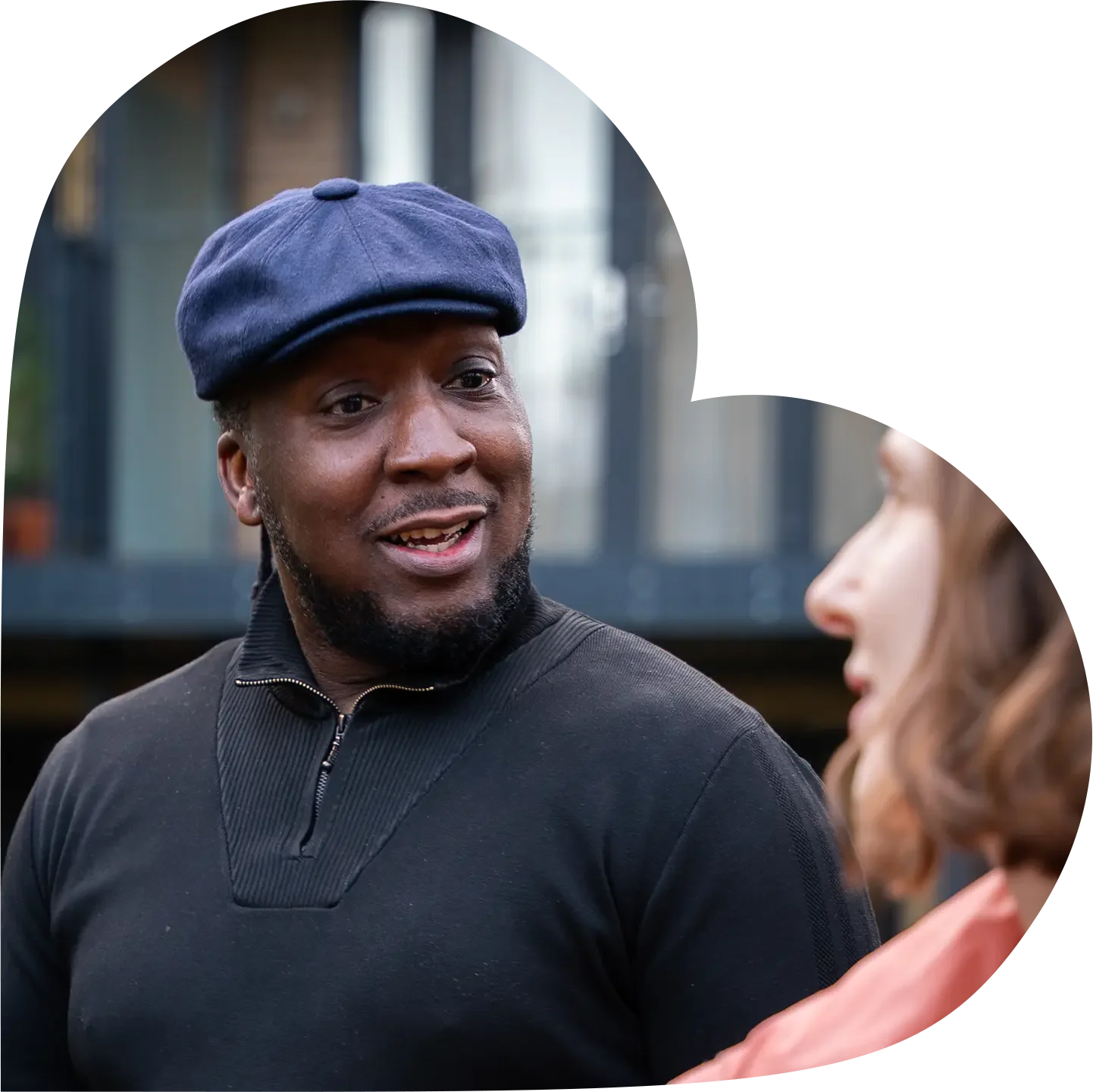 A man with a beard wearing a navy flat cap and a black zip-up sweater talking to a woman with shoulder-length brown hair and a pink shirt outdoors.
