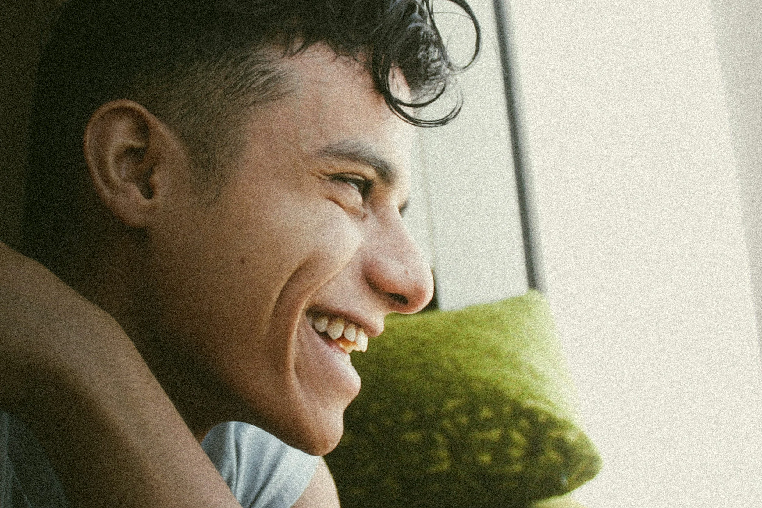 A young man with short, curly dark hair is smiling and looking out a window, with a green pillow behind his back.