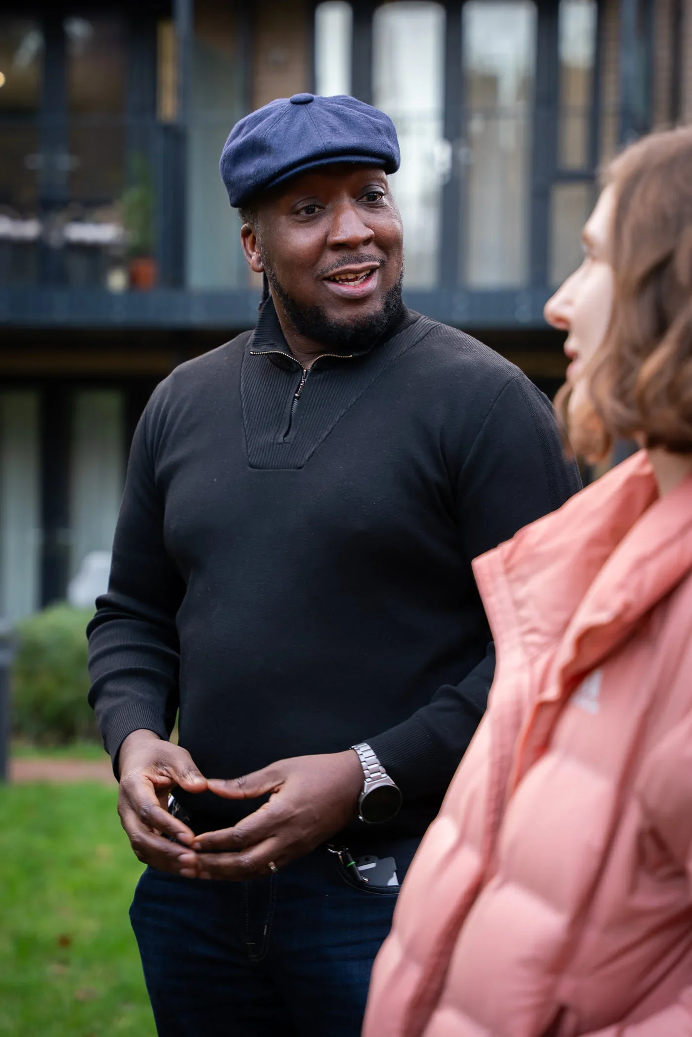 A man wearing a navy blue beret and black sweater is talking to a woman with brown hair and a peach-colored jacket outdoors.