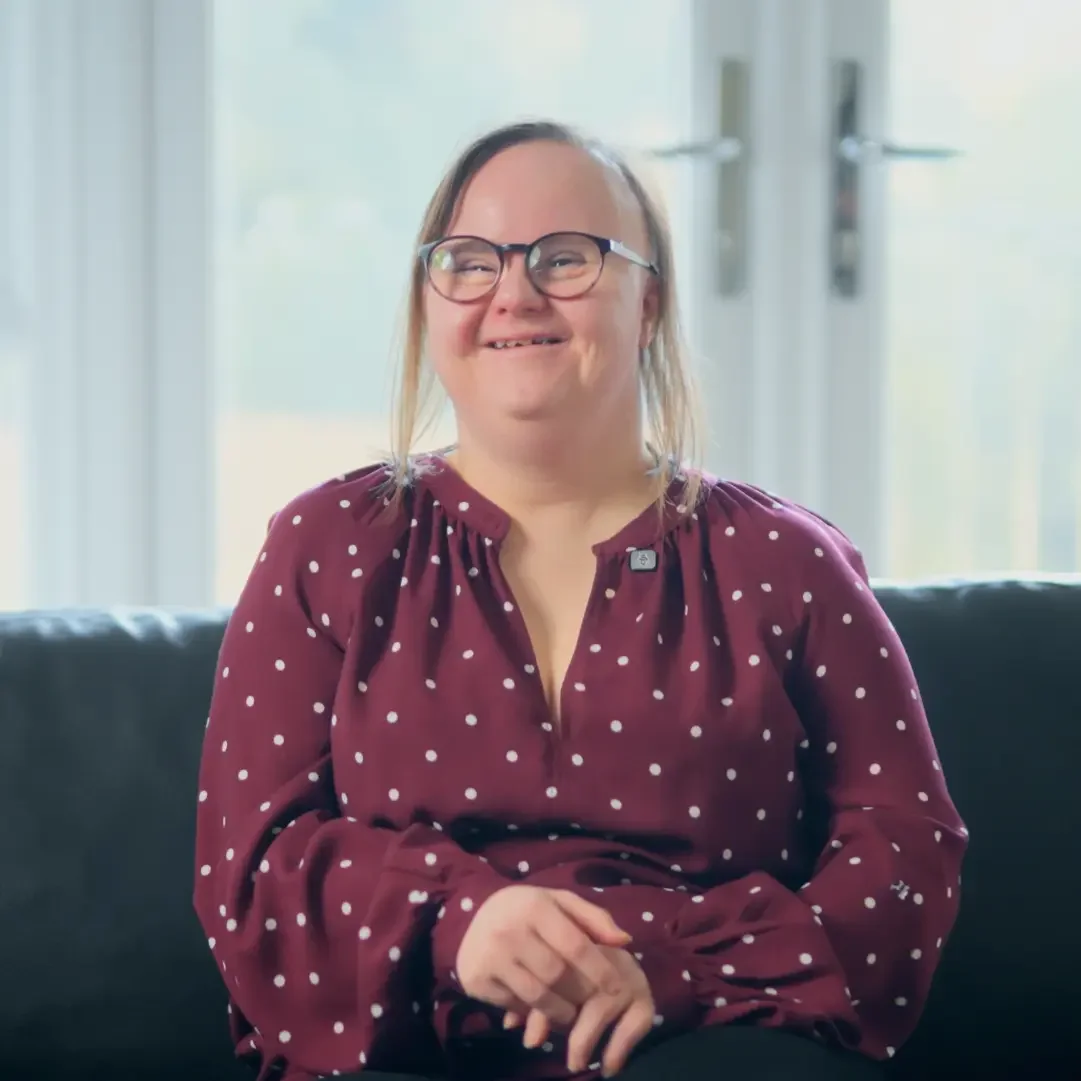 A woman with glasses and long hair, wearing a maroon polka dot blouse, smiling and sitting on a black couch in a bright room with large windows.