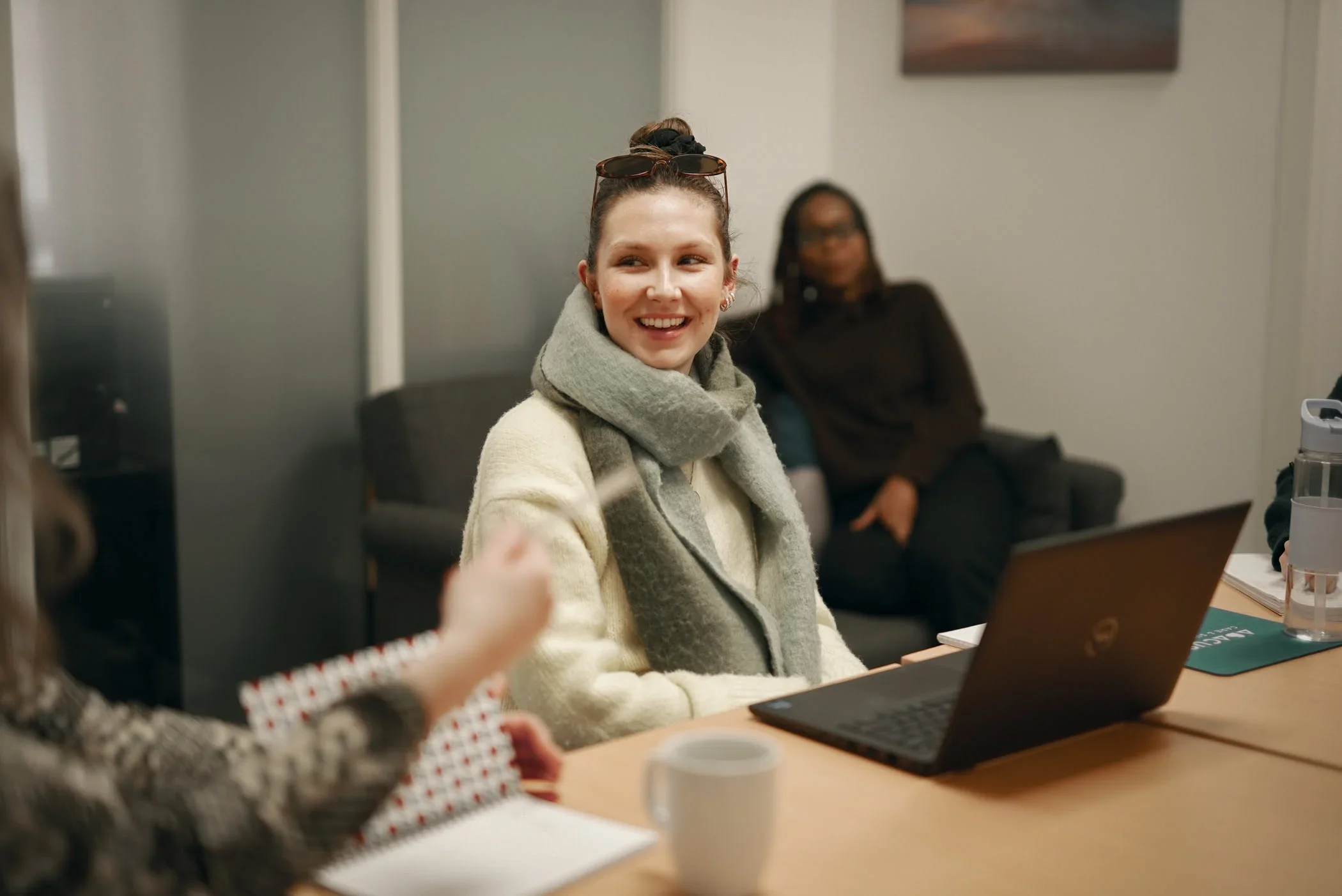 A woman with a scarf sitting at a conference table, smiling, with a laptop in front of her, in a meeting room with other women.