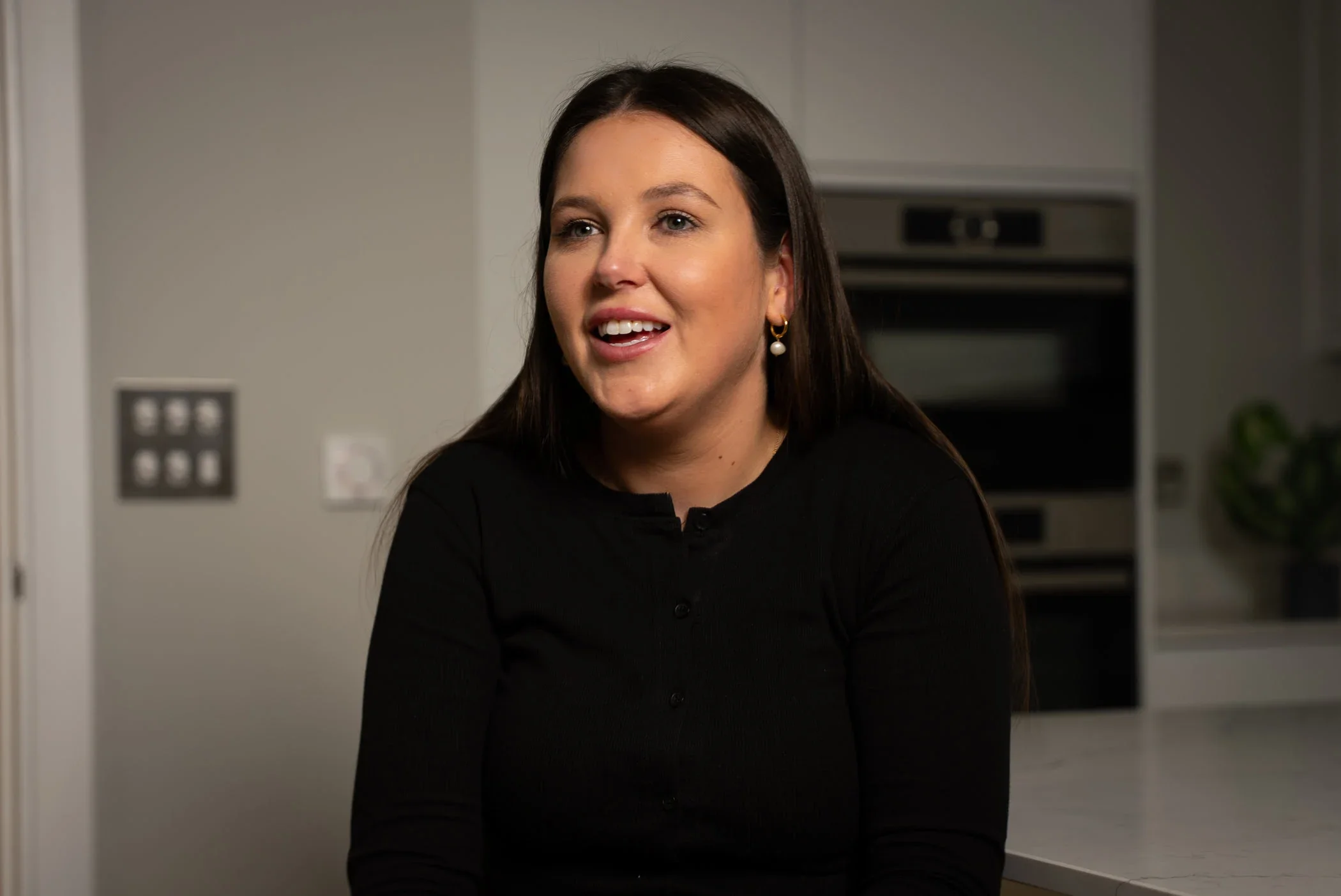 Woman with long dark hair, wearing a black top and pearl earrings, smiling in a modern kitchen.