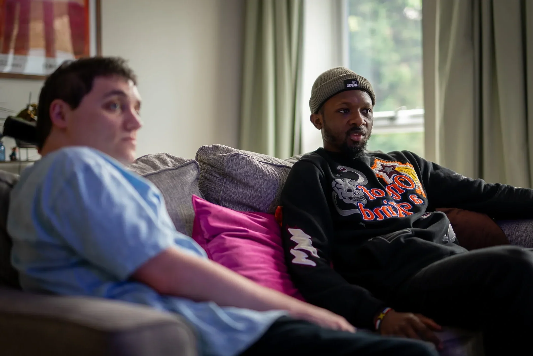 Two men sitting on a couch, one in a hospital gown and the other wearing a hoodie, with a window and curtains behind them.