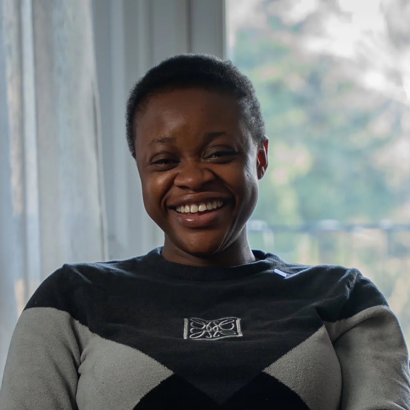 A woman with short hair smiling near a window with blinds, wearing a black and gray argyle sweater.