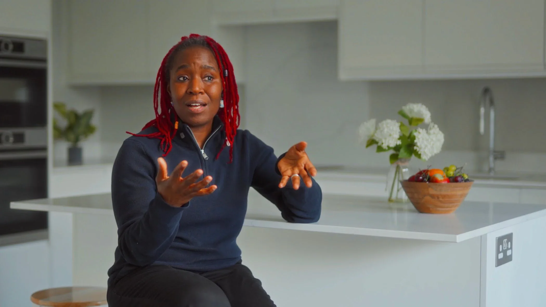 A woman with red dreadlocks gesturing while talking in a modern kitchen with white countertops and cabinets, a bowl of fruit, and white flowers.