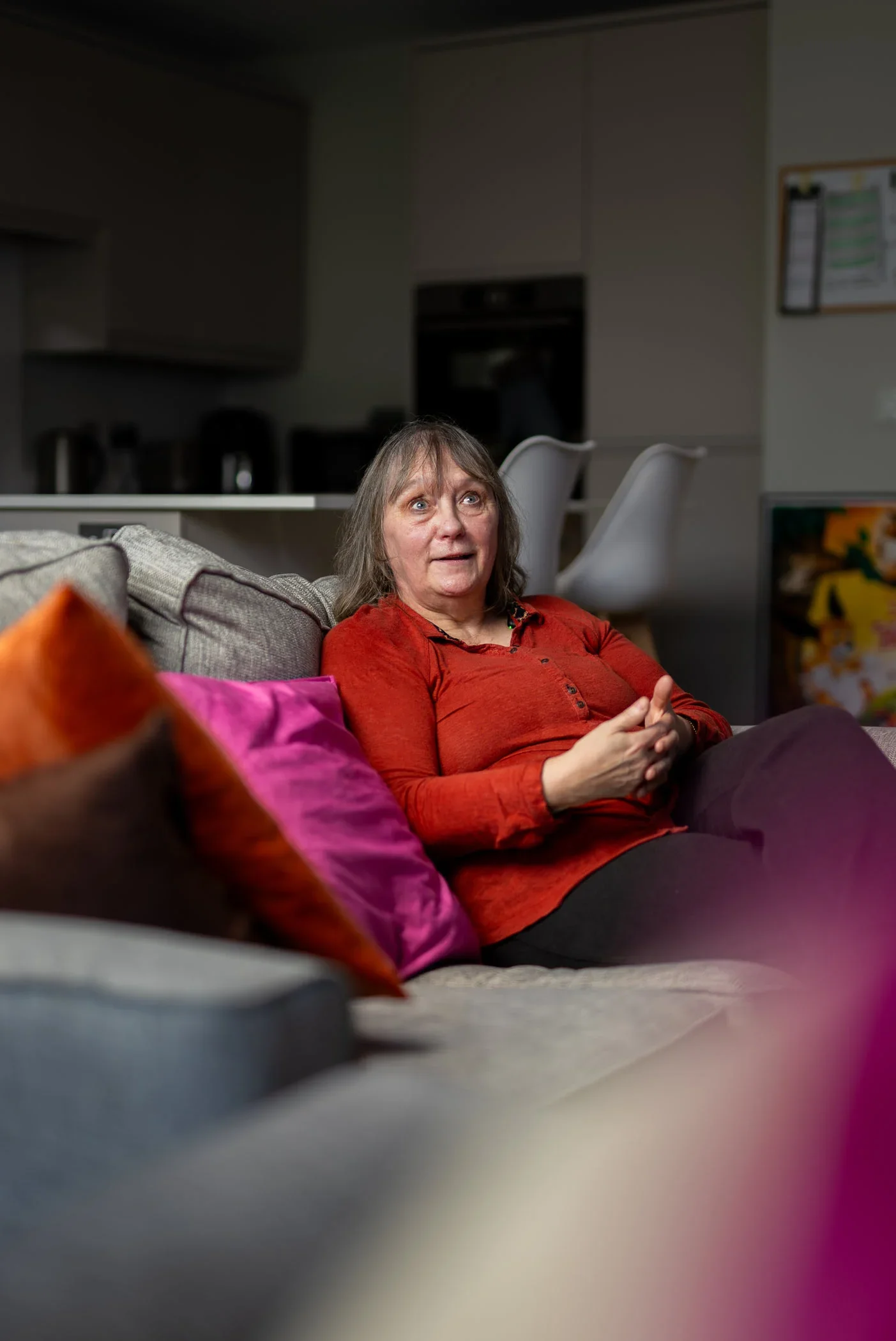 A woman sitting on a sofa with colorful pillows in a living room, appearing surprised or animated.