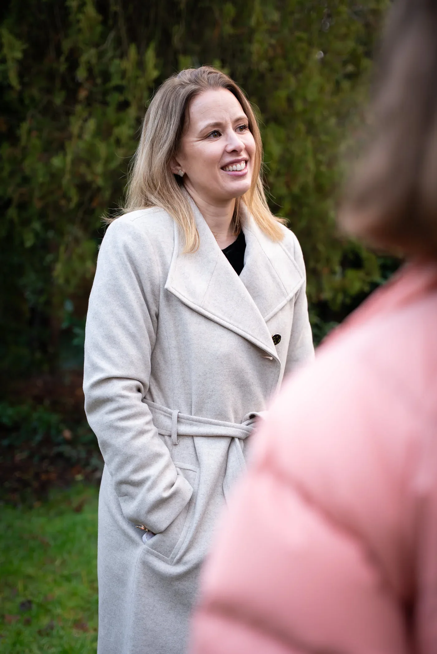 A woman with shoulder-length blonde hair smiling and wearing a beige coat standing outdoors in front of greenery.