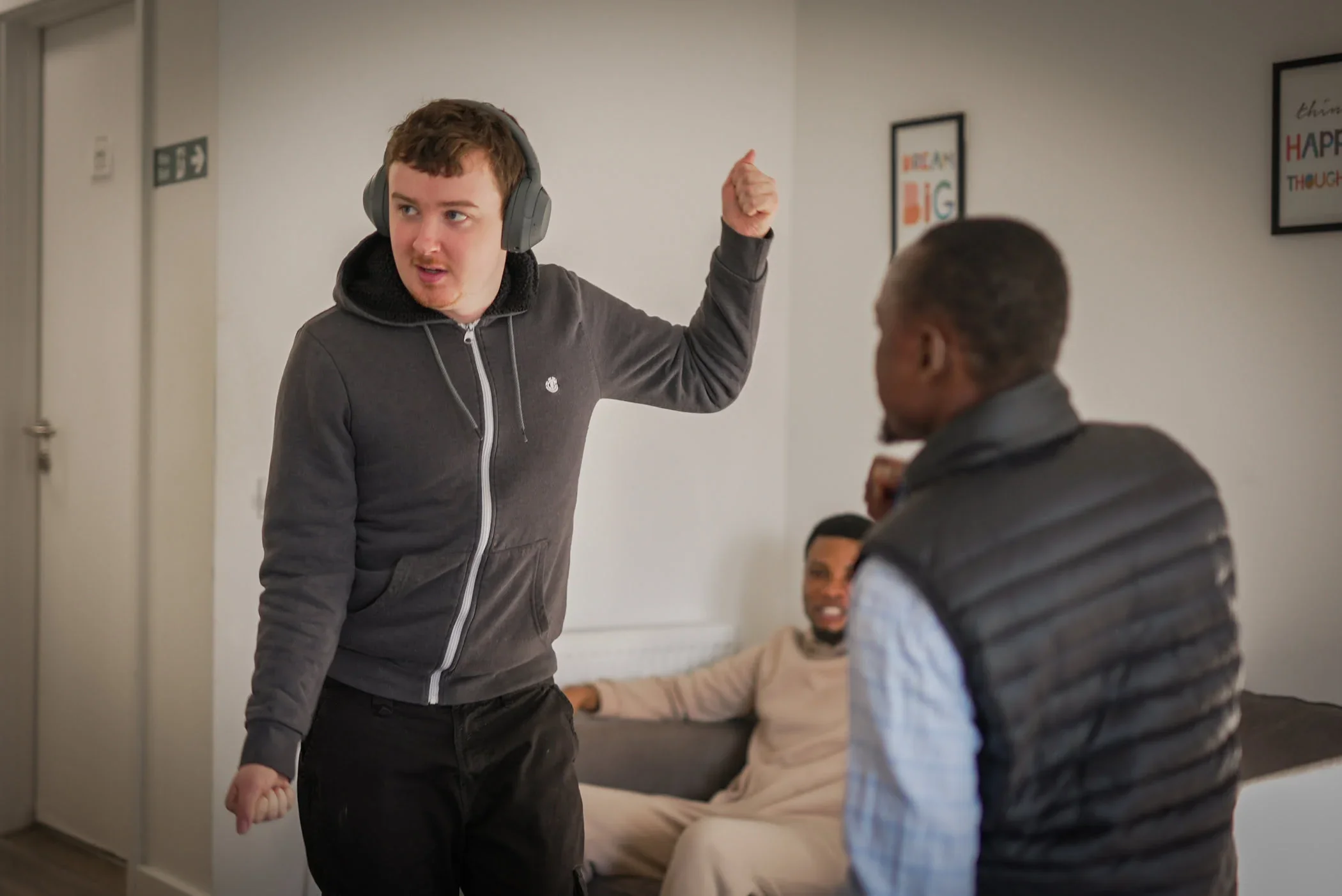 Three young men are inside a room, two of them are standing and engaged in a conversation, while the third is sitting on a sofa in the background. The man in the foreground on the left is wearing headphones, a gray hoodie, and appears to be speaking or arguing with the man in the foreground on the right, who is wearing a black padded vest. The man in the background is sitting comfortably on a sofa and smiling.