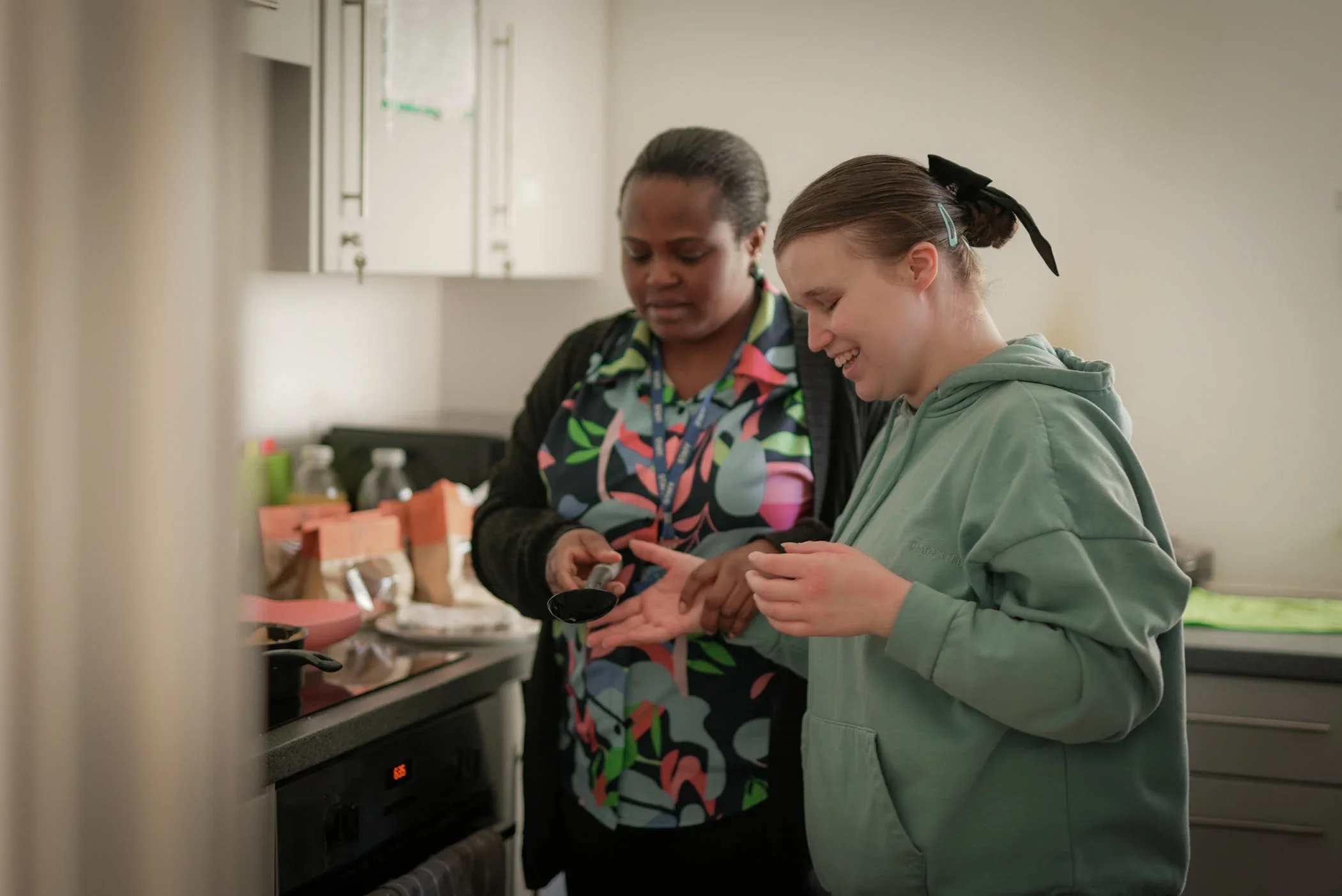 Two women are in a kitchen, one is pouring seasoning into her hand while the other watches and smiles.