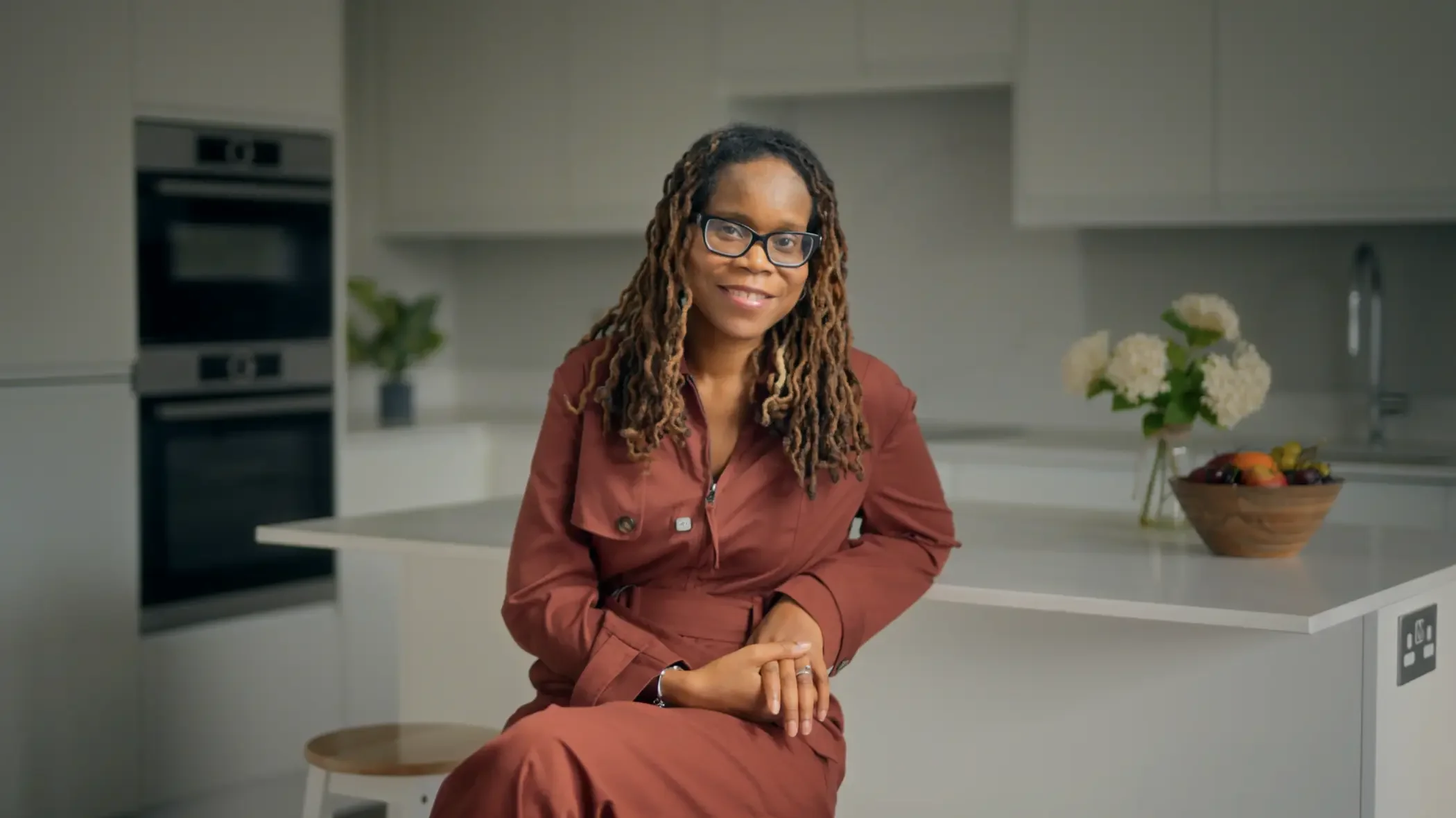 A woman with glasses and dreadlocks sitting on a stool in a modern kitchen, smiling at the camera with a bowl of fruit and a vase of white flowers on the counter behind her.