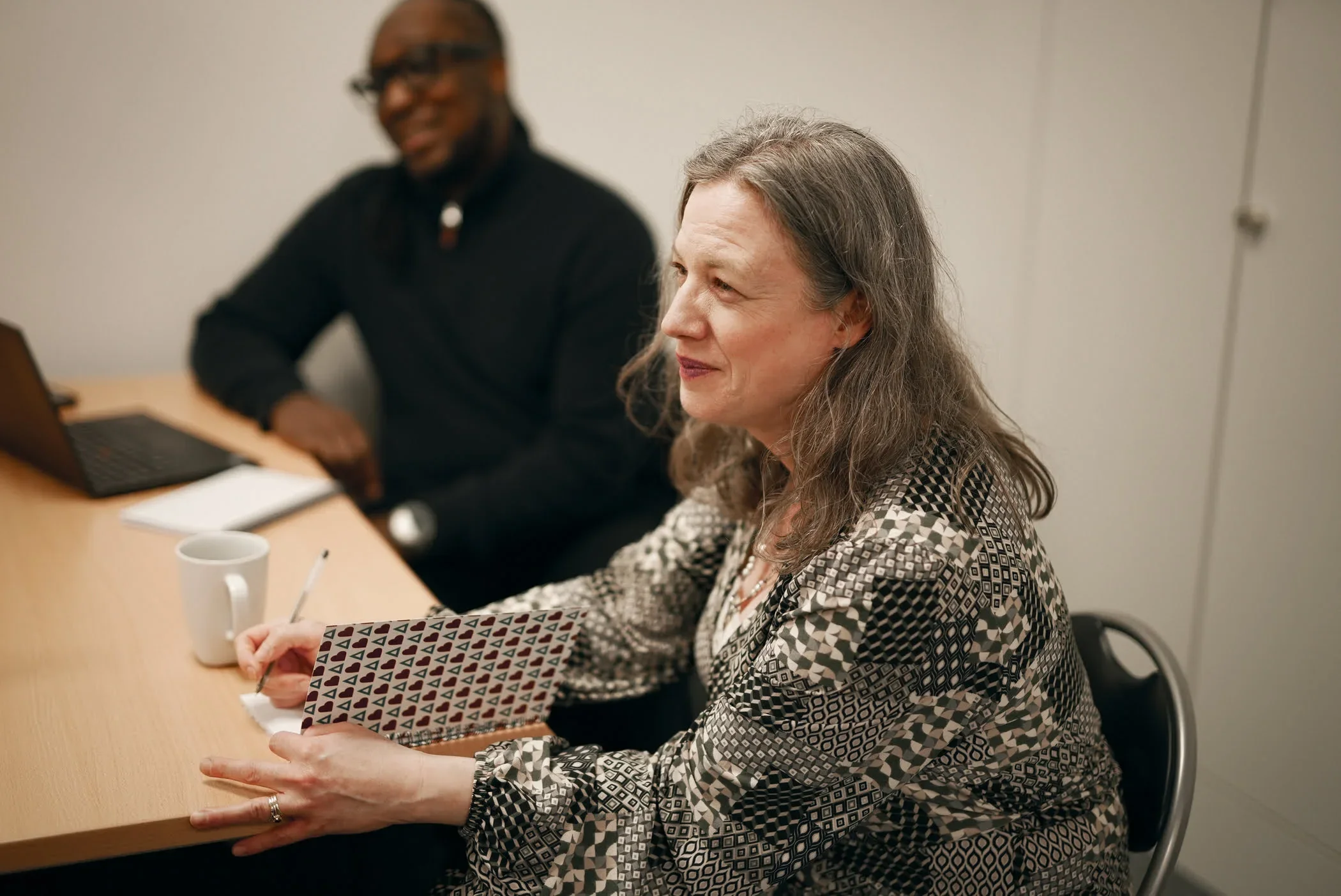 A woman with gray hair sitting at a conference table, holding a notebook, with a mug and pen in front of her, during a meeting with a man in the background.