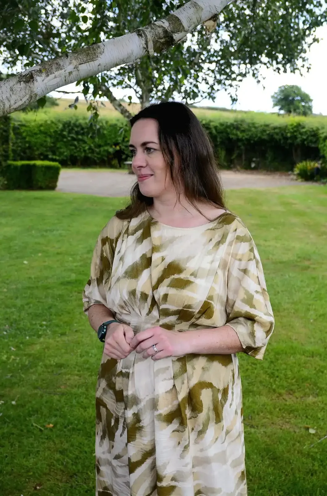 A woman with shoulder-length brown hair smiling and wearing a green dress tanding outdoors in front of greenery.
