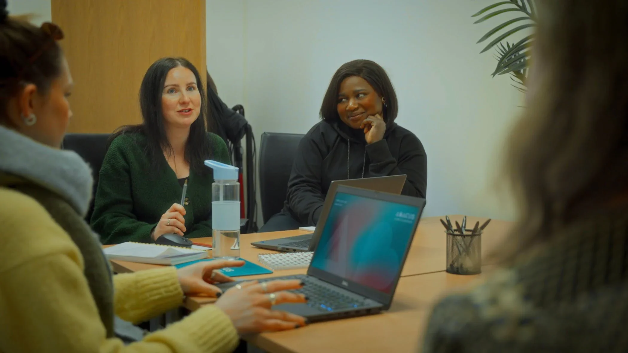 Women in a meeting room, sitting at a table with laptops, notebooks, pens, and a spray bottle, engaged in conversation.